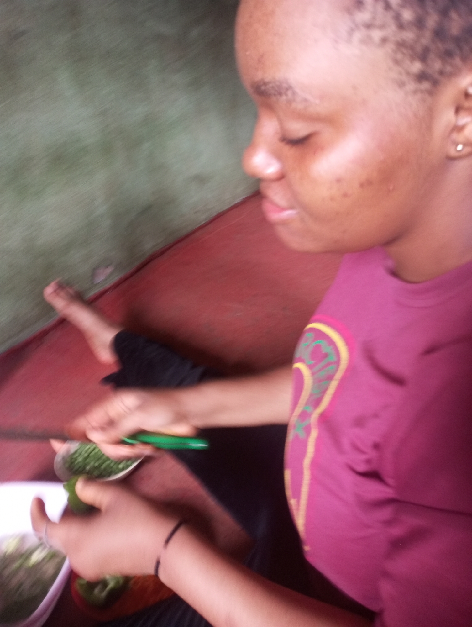 A medium shot shows a young woman with short hair, wearing a maroon t-shirt with a yellow and green trim on the left side, preparing food. She is seated, with her left foot bare and resting on a red surface. Her hands are blurred as she cuts green vegetables with a green knife, holding a white bowl and a red cutting board. The background shows a textured wall, part green and part a darker color, and a red floor. The scene appears to be indoors. This image was captured in Onitsha, Nigeria.