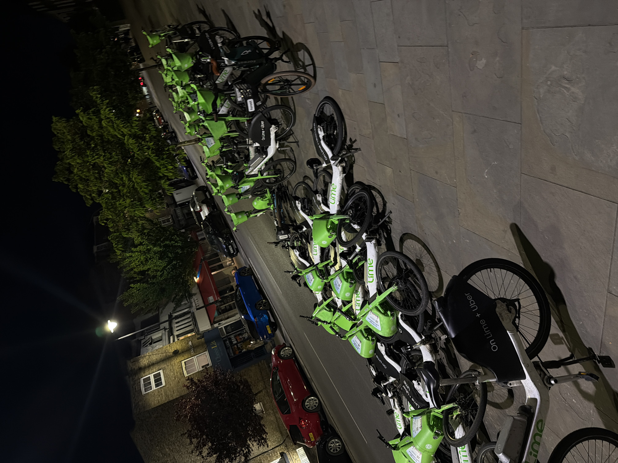 The image captures a nighttime street scene in London, United Kingdom. A long, orderly row of bright green and white Lime bicycles and electric scooters dominates the foreground, parked along a paved sidewalk. Many of these shared mobility vehicles display "Lime" branding, with additional text such as "On Lime + Uber" visible on their seats and frames. One black bicycle from a different service, labeled "forest," is also present among the Lime vehicles.
Adjacent to the sidewalk, a street features several parked cars, including a distinct blue vehicle, a red car, and other darker automobiles. Across the street, various buildings are visible, one featuring a brick facade with white-framed windows. Trees and bushes line the street, adding greenery to the urban environment. The scene is illuminated by streetlights and light emanating from building windows, casting shadows and highlighting the vibrant green of the bikes against the dark sky. The pavement is composed of large, rectangular stone slabs.
Visible commercial signs include "ChocoLit," "BARBERS," and a partially obscured "BEAR." The time of day is clearly night, indicated by the dark sky and artificial lighting. No people are visible, and the weather appears dry.