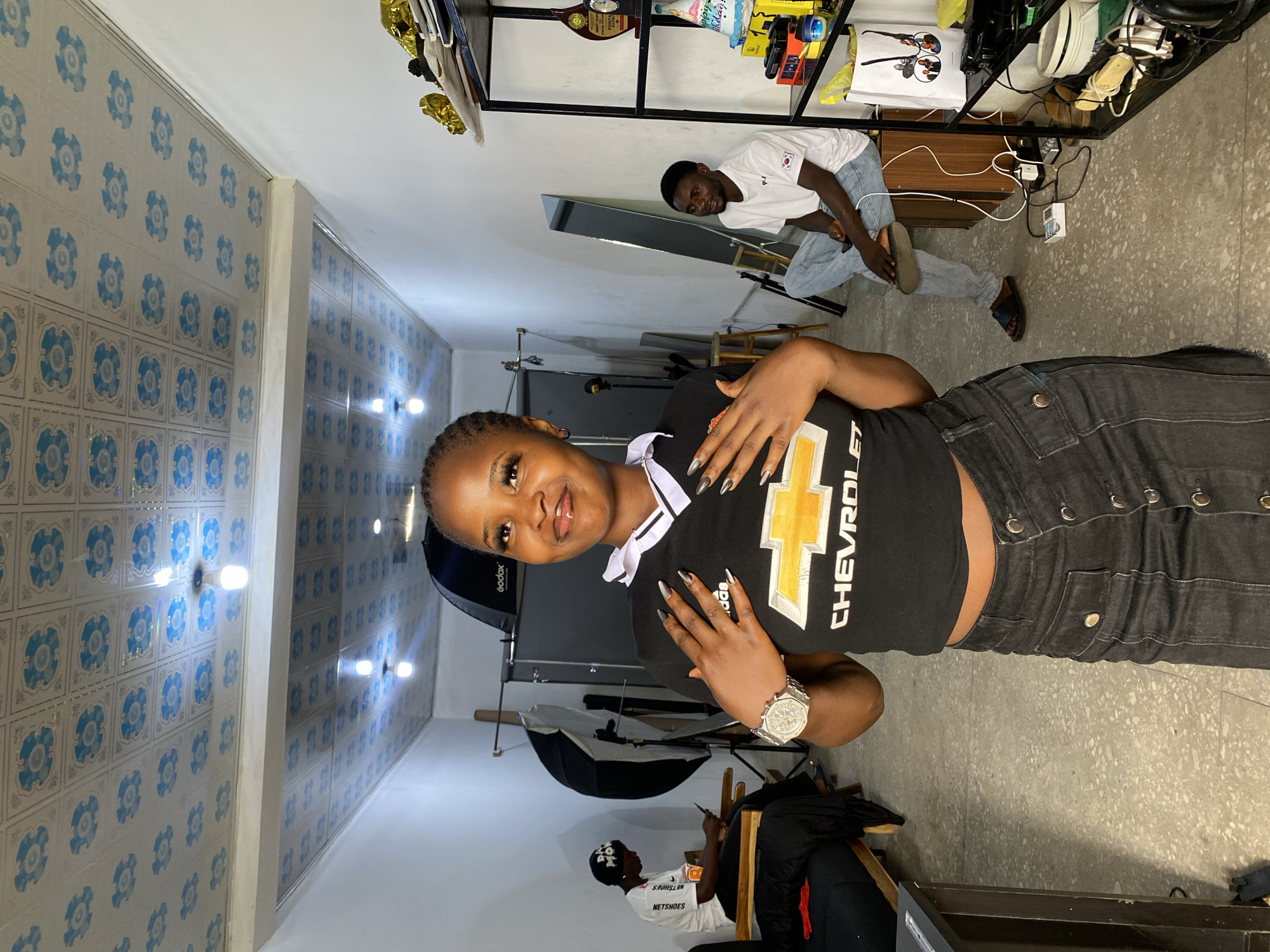 In Gura Top, Nigeria, a young woman stands in a studio space, posing with her hands on her chest. She wears a black crop top with a white collar and a Chevrolet logo, paired with a black denim skirt. A large, silver watch adorns her left wrist. In the background, a man sits on a wooden stool, looking at his phone, wearing a white t-shirt and light blue jeans. Another person, wearing a black beanie and white t-shirt, is seated on a couch to the left. The studio features photography equipment, including softboxes and umbrellas, and shelves displaying various items. The ceiling is covered with decorative tiles.