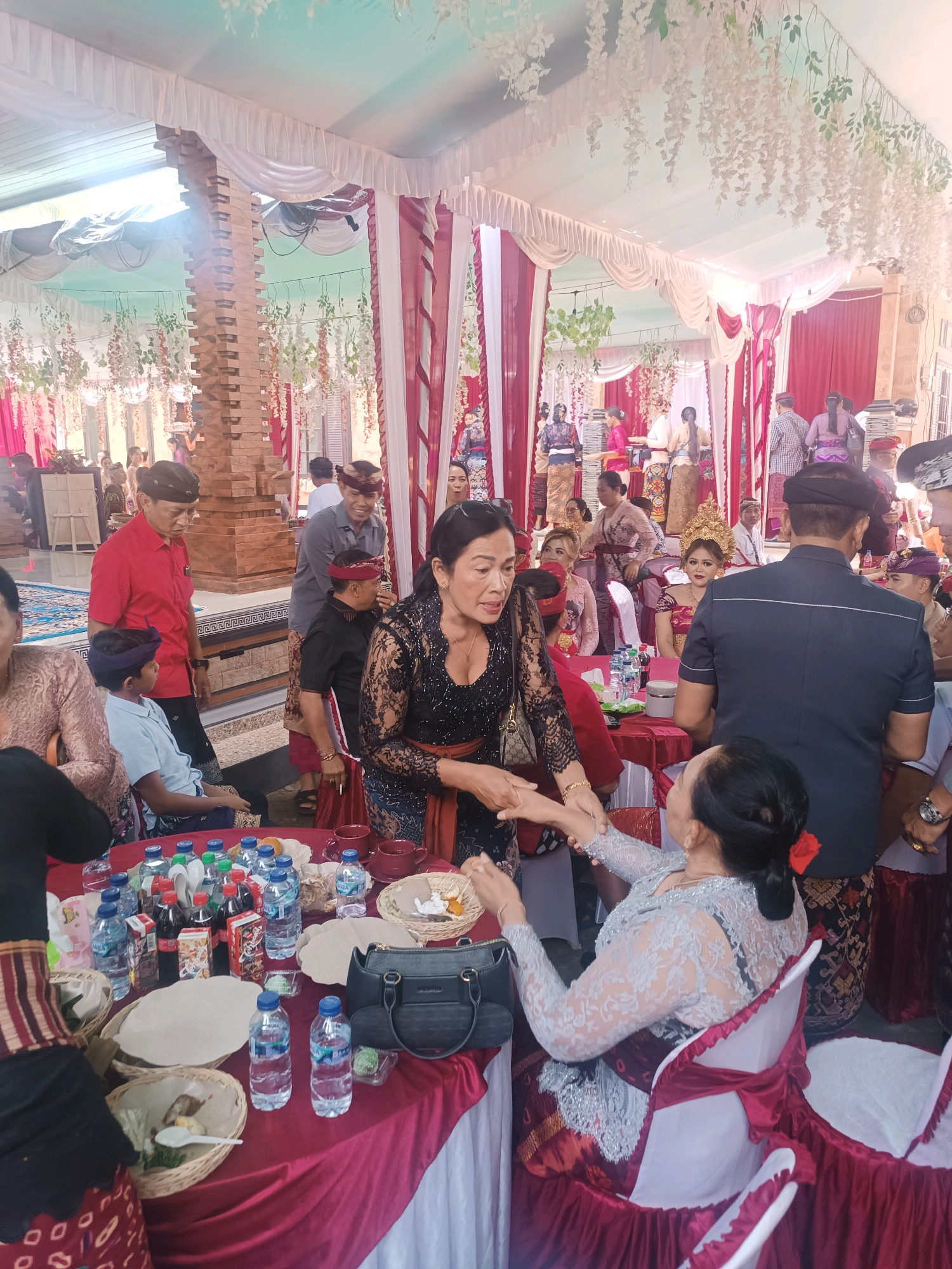 In Penebel, Indonesia, a group of people are gathered at an indoor event, possibly a wedding or cultural ceremony. The environment is decorated with white and red draped fabric and white floral garlands hanging from the ceiling. A brick pillar is visible on the left side of the frame. A long, red-covered table is laden with drinks, food items in woven baskets, and bags. Several individuals are dressed in traditional Indonesian attire, including sarongs and elaborate tops.
In the foreground, a woman in a black lace top is interacting with another woman in a grey lace dress who is seated. The woman in the black top is holding out her hand, and the seated woman is reaching for it. To the left, a child in a blue shirt and a purple sash is seated. A man in a red shirt and a man in a grey shirt with a purple sash are standing behind the seated child.
In the middle ground, more people are visible, including a woman in a reddish-pink traditional outfit and a golden headdress. Several other guests are seated and standing, with many wearing traditional clothing. The overall atmosphere appears to be one of celebration and community.