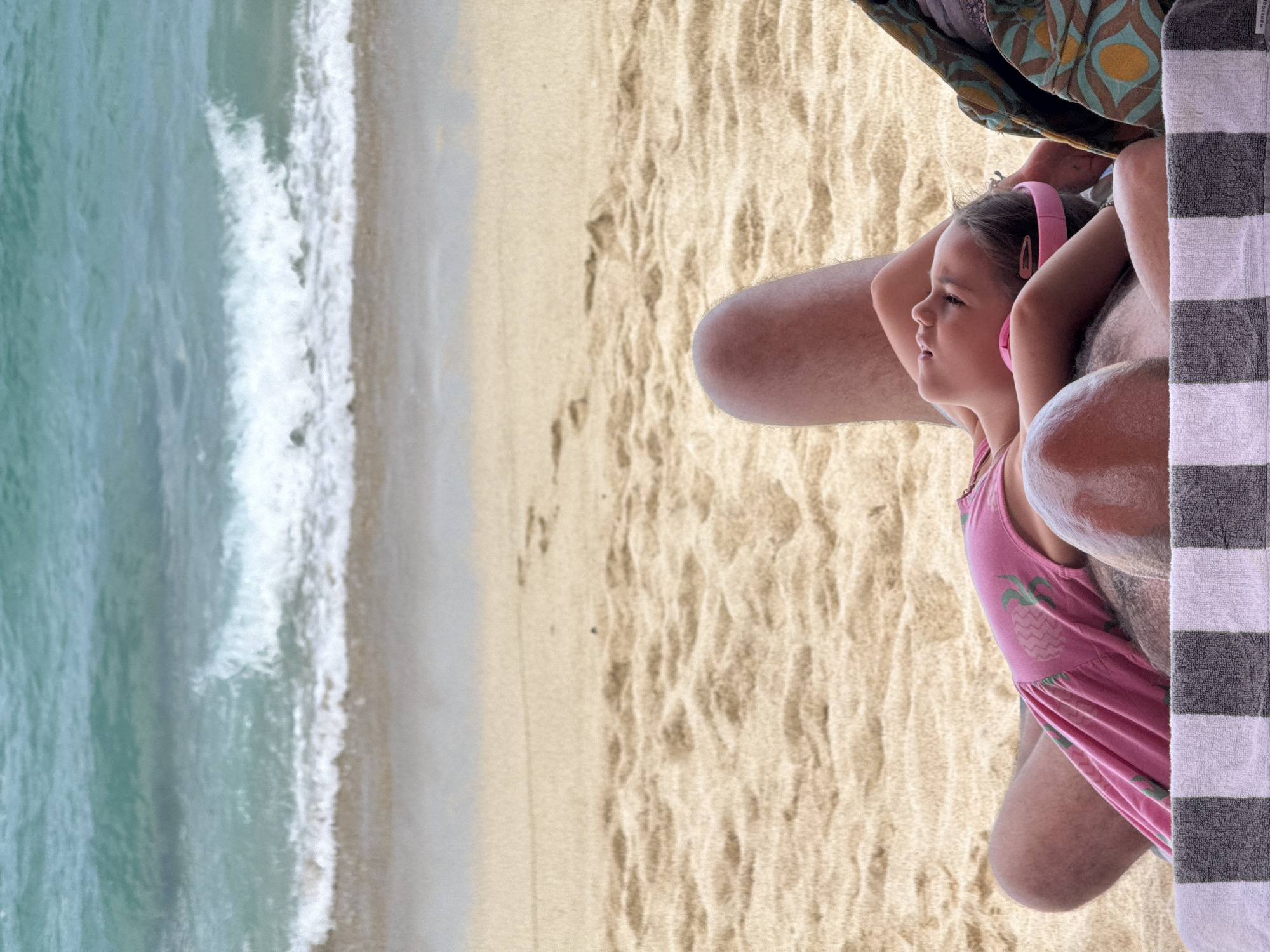 A medium-shot, eye-level photograph depicts a child with pale skin, wearing a pink dress with pineapple designs and pink headphones, seated on the sandy beach of Kuta Selatan, Indonesia. The child is positioned to the right side of the frame, and their gaze is directed towards the left of the frame, where the ocean meets the sandy shore. A body of water with white waves is visible in the background on the left. The beach sand is light brown. Parts of an adult’s body (legs and arms) are visible, as is a patterned blanket and a vertically-striped towel.