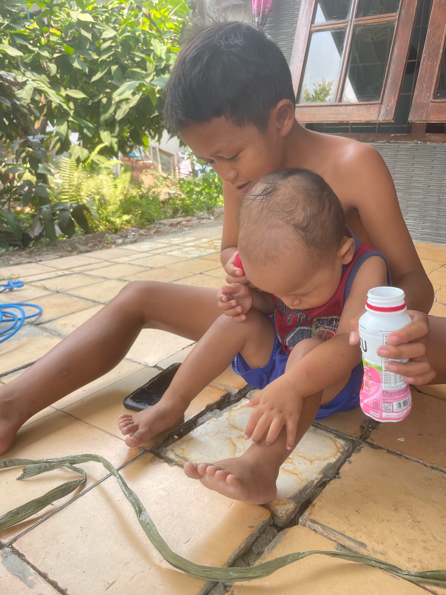 This media file depicts two young children, likely siblings, sitting on an outdoor tiled patio in what appears to be a residential setting in Kebon Waris, Indonesia. The older child, a boy with dark hair, is positioned behind the younger child, a baby or toddler. The older child is shirtless and is looking down towards the younger child, his arm gently touching the younger child's leg. The younger child is wearing a red and blue sleeveless shirt and blue shorts. The younger child is looking downwards, and its hand is near its mouth.
In front of the children, on the patio, are several objects. A black smartphone is lying on the ground. To the right of the younger child, a white bottle with a pink label is held by the older child. The label on the bottle appears to have text and graphics, but it is partially obscured. A bright blue coiled rope lies on the patio to the left. A long, thin, dry green leaf or reed is also present on the patio, winding across the foreground.
Behind the children, lush green foliage and trees are visible, suggesting an outdoor environment. A wooden-framed window with glass panes is also present in the background, hinting at a building. The lighting suggests it is daytime, likely with bright sunlight given the shadows and the overall illumination. The scene conveys a sense of casual interaction between the children in a warm, possibly tropical, climate.