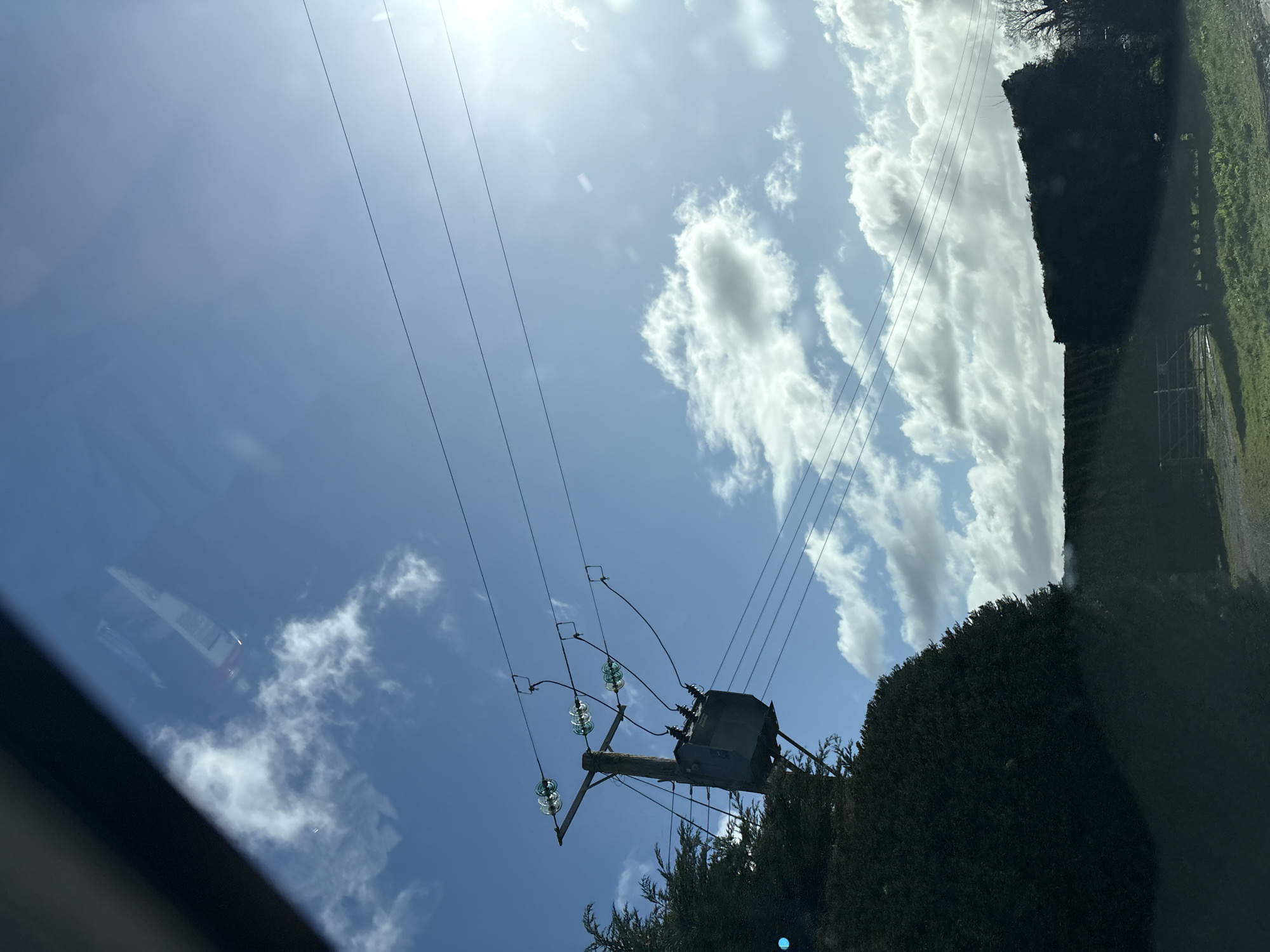 The image presents a daytime outdoor scene in Ruyton-XI-Towns, United Kingdom, captured from inside a vehicle, indicated by reflections in the upper left. A wooden utility pole stands prominently on the left, supporting a grey, cylindrical electrical transformer. Several green-tinted glass insulators are visible on the pole, connecting the transformer to three parallel overhead power lines stretching across the upper frame. Dark green, dense evergreen hedges and trees occupy the lower left and extend across the middle ground. In the lower right, a patch of green grass is visible, partially obscured by a lower hedge, behind which a metal gate and a dirt or gravel track are discernible. A bare deciduous tree is visible against the sky on the far right. The sky is clear blue with scattered white cumulus clouds, and the bright sun is present in the upper right, causing a lens flare effect. No individuals or active interactions are observed in the scene.
