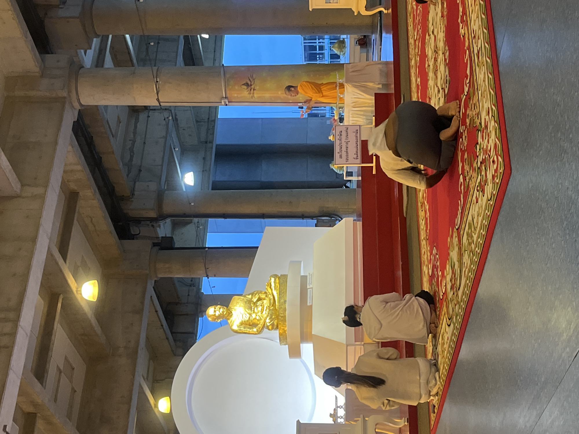 In Tha Khlong Town Municipality, Thailand, a Buddhist ceremony is underway. A golden statue of a seated monk, likely a revered figure, is prominently displayed on a white tiered platform. In front of the statue, three individuals are kneeling on a red carpet adorned with intricate patterns. Two women, facing away from the camera and towards the statue, are dressed in light-colored attire. A third individual, also kneeling and facing away, wears a white head covering and dark clothing. To the right, a monk in saffron robes stands at a table, holding an object and appearing to lead the ceremony. A sign with Thai script is placed on the table. The environment features large concrete pillars and a textured ceiling, illuminated by overhead lights, suggesting an indoor or covered outdoor space. The overall scene depicts a religious gathering with elements of reverence and tradition.