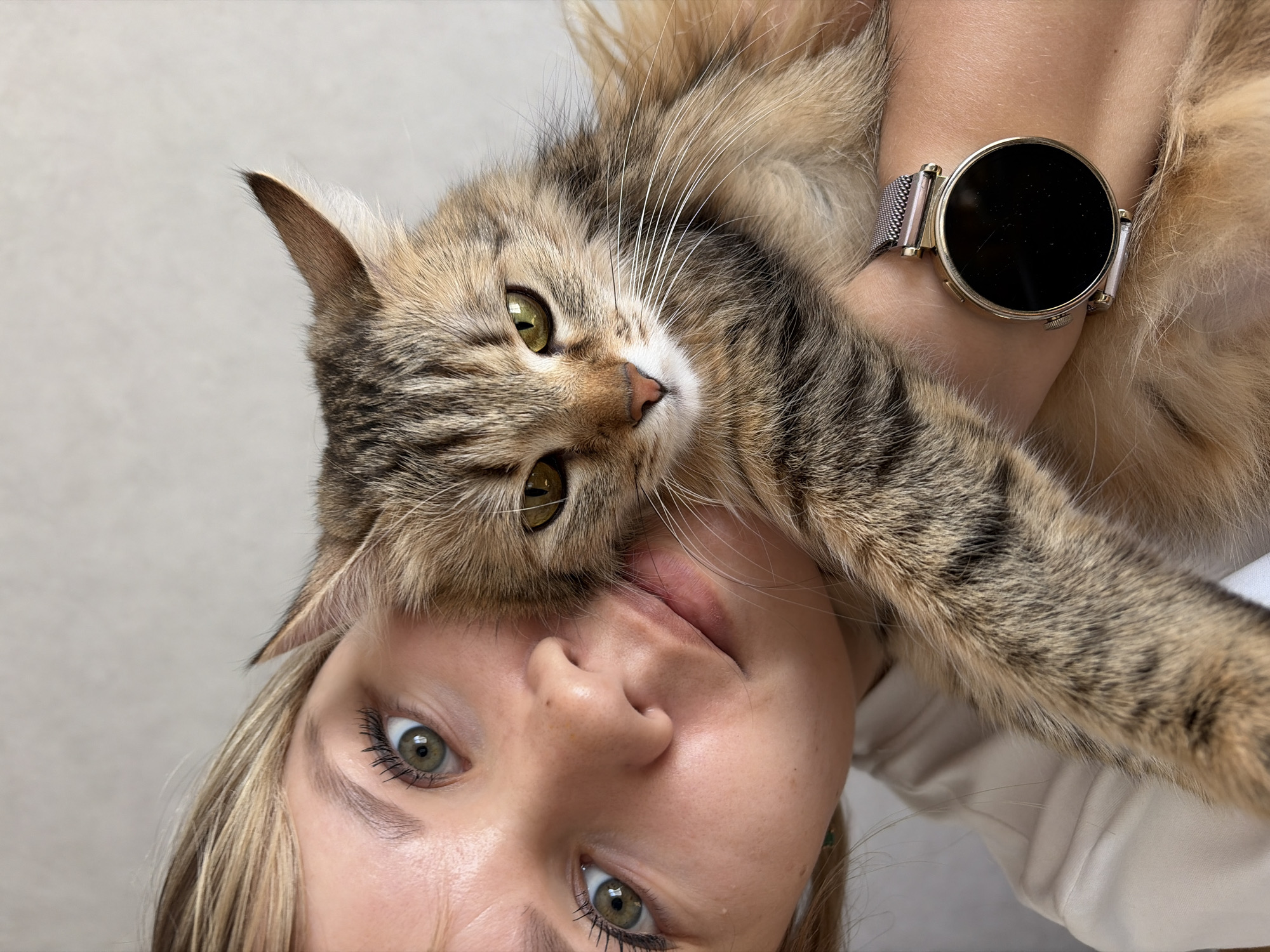 A selfie depicts a person holding a tabby cat. The person has fair skin, green eyes, and blonde hair with bangs. They are wearing a gold smart watch. The cat has green eyes. The location is Chaikovsky, Russia.