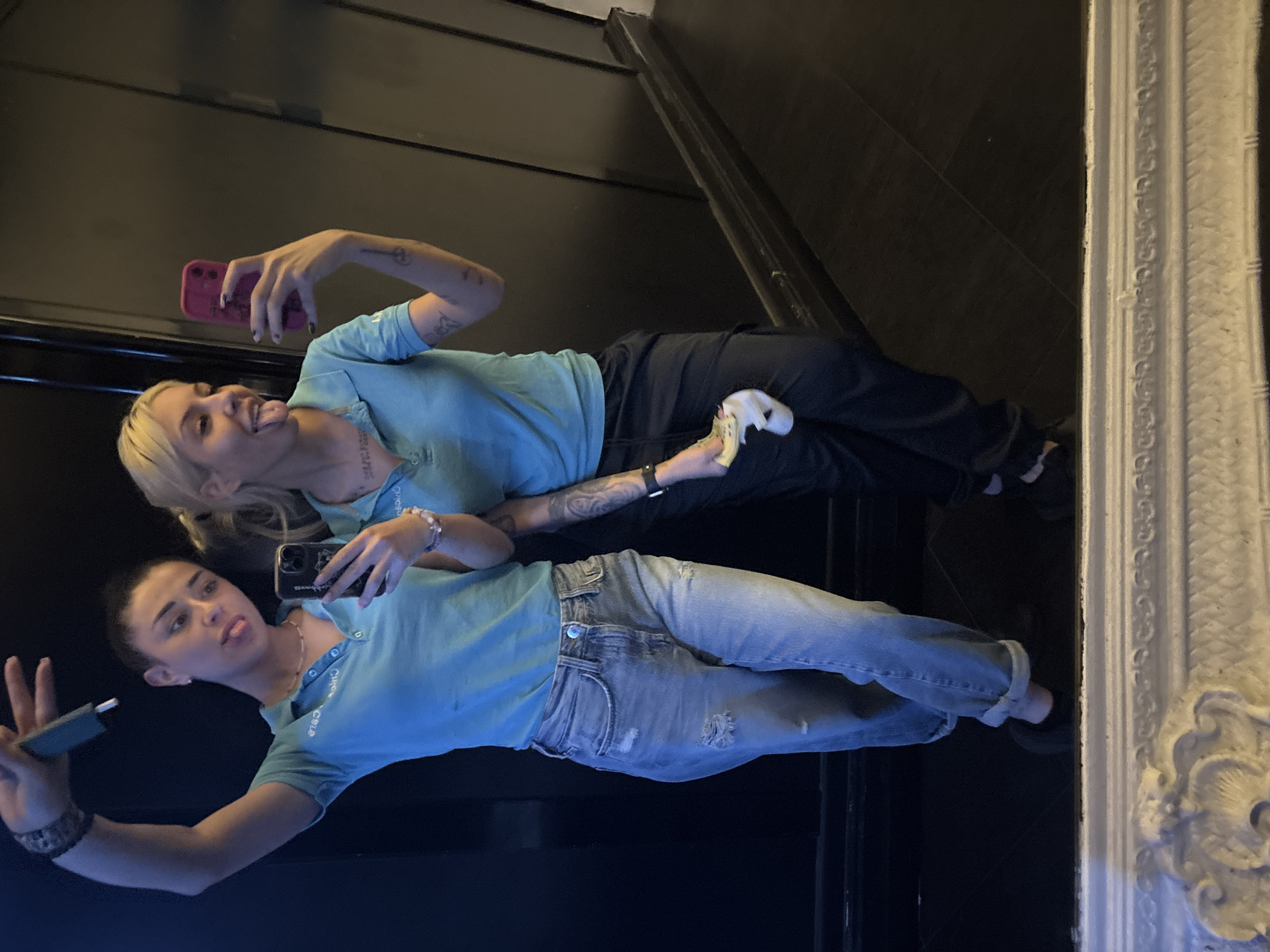 Two young women are reflected in a mirror. The woman on the left is holding a phone and appears to be taking a selfie. She is wearing a blue polo shirt and light-wash jeans. She has short dark hair and a bracelet. The woman on the right is also holding a phone and sticking her tongue out. She is wearing a similar blue polo shirt and dark pants. She has blonde hair. She appears to be holding money or receipts in her left hand. The background appears to be a dark hallway or room with a decorative white mirror frame visible on the right side of the image. It is not possible to ascertain the specific location in Plovdiv, Bulgaria from this image alone.