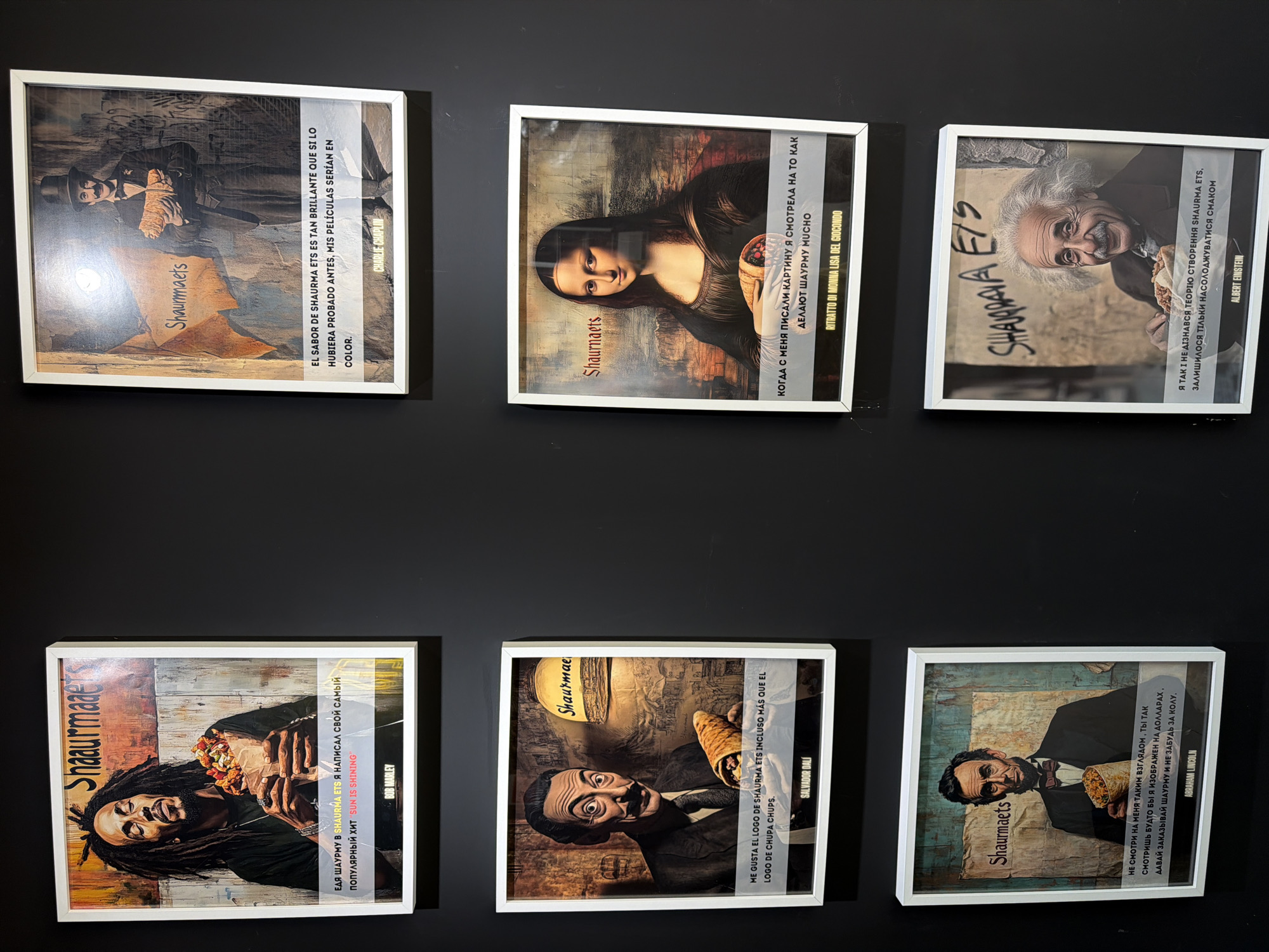 The image shows six framed posters hanging on a dark wall. Each poster features a famous person holding a shawarma and a text related to shawarma and their identity. The posters depict Charlie Chaplin, Mona Lisa, Albert Einstein, Bob Marley, Salvador Dalí, and Abraham Lincoln. The word "Shaurmaets" or a similar variant is present on each poster, presumably as a brand name. The setting is likely an interior space. The posters appear to be promotional materials or artistic decorations. Given the word "Shaurma", it might be inside a restaurant specializing in shawarma. The image doesn't provide explicit clues about the city Valencia in Spain, but this might be inferred if there's additional contextual information about this Shawarma restaurant.