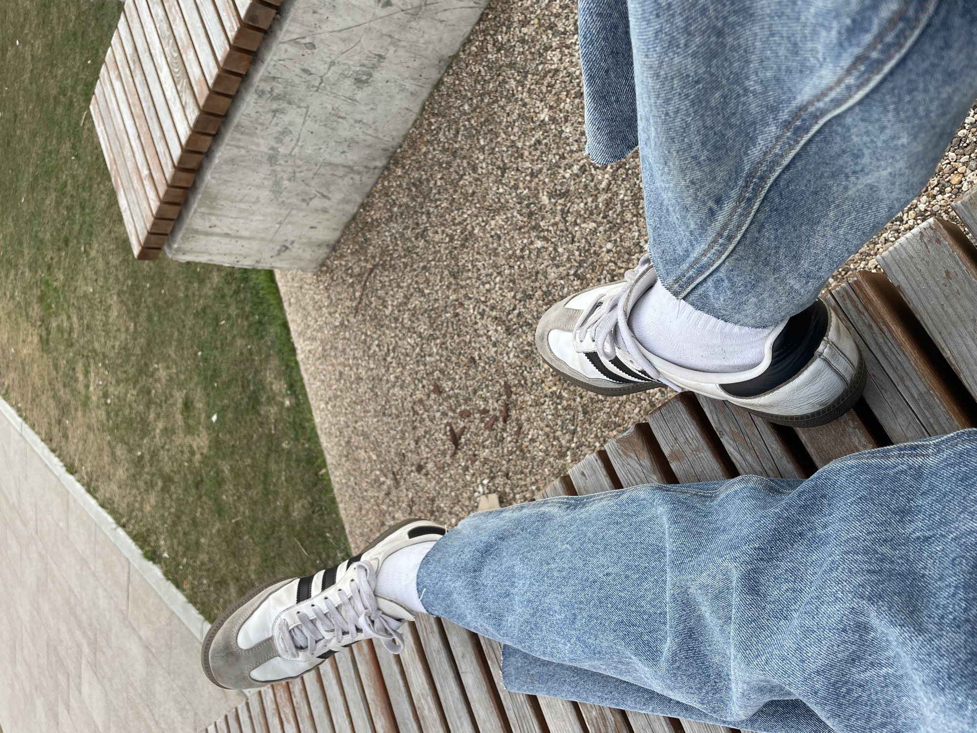 The image shows a first-person view of someone sitting on a wooden bench in an outdoor setting. The person is wearing light blue jeans and white Adidas Samba sneakers with white socks. The bench has a slatted design. Adjacent to the bench is a paved area with small pebbles, and beyond that is a patch of green grass and another concrete bench. The photo does not contain information about the location in the city of Moscow, Russia.