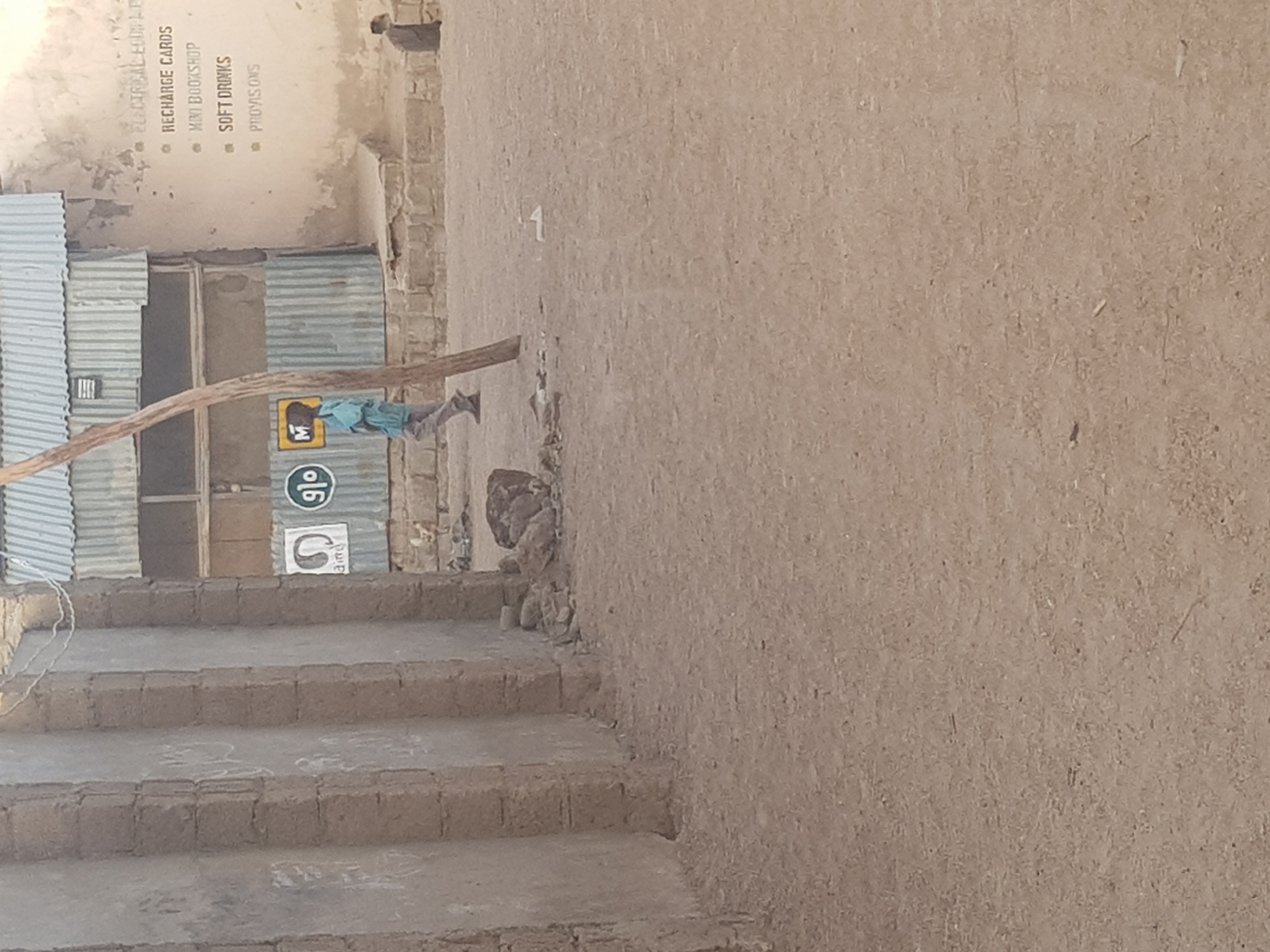 The image depicts a street scene in Dutsinma B, Nigeria. On the left, there are a set of concrete stairs leading up to a building with a corrugated metal facade. Painted on the wall are advertisements for "Glo" and "airtel," along with a sign listing "Recharge Cards," "Mini Bookshop," "Soft Drinks," and "Provisions." A wooden branch extends diagonally across the frame. A person wearing blue clothing is walking near the entrance. The background features a tan-colored wall.