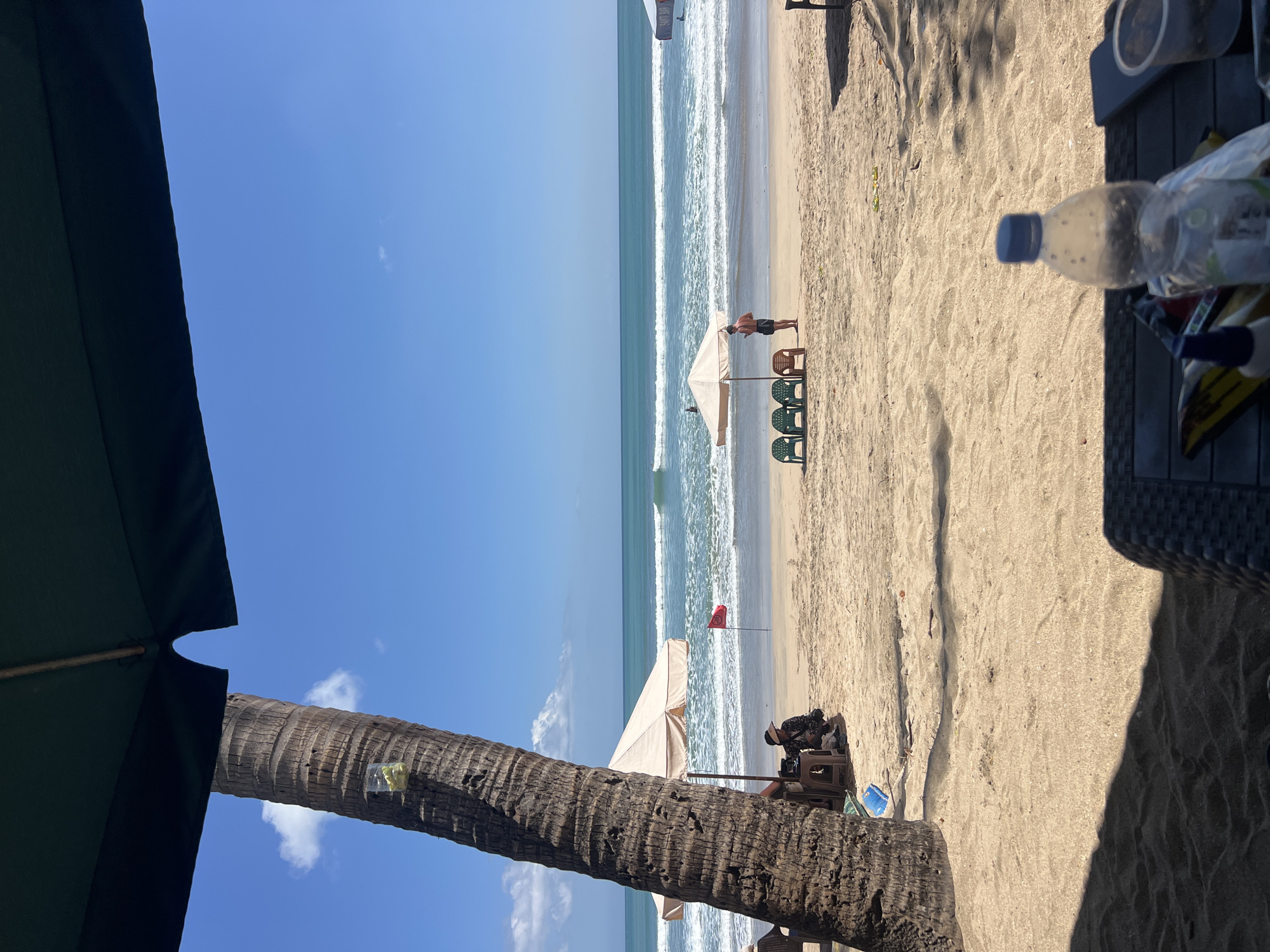 The image depicts a serene beach scene in Kuta, Indonesia. The foreground is dominated by the rough texture of a palm tree trunk and the shadow of a dark umbrella, hinting at a shaded seating area. Beyond, the golden sand stretches to meet the turquoise ocean, where gentle waves break. Several beach umbrellas, some beige and one white, are scattered along the shoreline, providing shade. A few plastic chairs, colored green and brown, are arranged near one of the umbrellas.
A few individuals are visible. One person, wearing a hat and dark clothing, is seated under a beige umbrella, appearing to be resting. Another person, a man in shorts, stands near a cluster of chairs and an umbrella, looking out at the sea. A red flag planted in the sand suggests a designated swimming area or a warning. Further out in the water, faint figures of people can be seen enjoying the ocean. The sky is a vibrant, clear blue with a few wispy clouds. In the immediate foreground, on a table, a plastic water bottle and some snack packaging are visible. The overall impression is one of a tranquil day at a popular tourist beach.