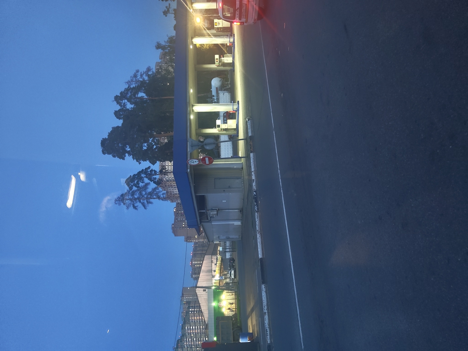 The image captures a nighttime or twilight view of a gas station in Ramenskoye, Russia. The gas station features a blue-roofed canopy illuminated by bright lights, supported by white pillars. Several fuel pumps are visible under the canopy, with a large white cylindrical tank behind them. A white car with its brake lights on is positioned to the right of the scene, and a smaller red vehicle is parked under the canopy. To the left, a gray building is attached to the gas station structure. In the background, there are residential buildings with illuminated windows, and tall evergreen trees are silhouetted against the dark blue sky. A road with a white dividing line occupies the foreground. The overall impression is one of a functional, well-lit commercial area in a Russian city at dusk.
