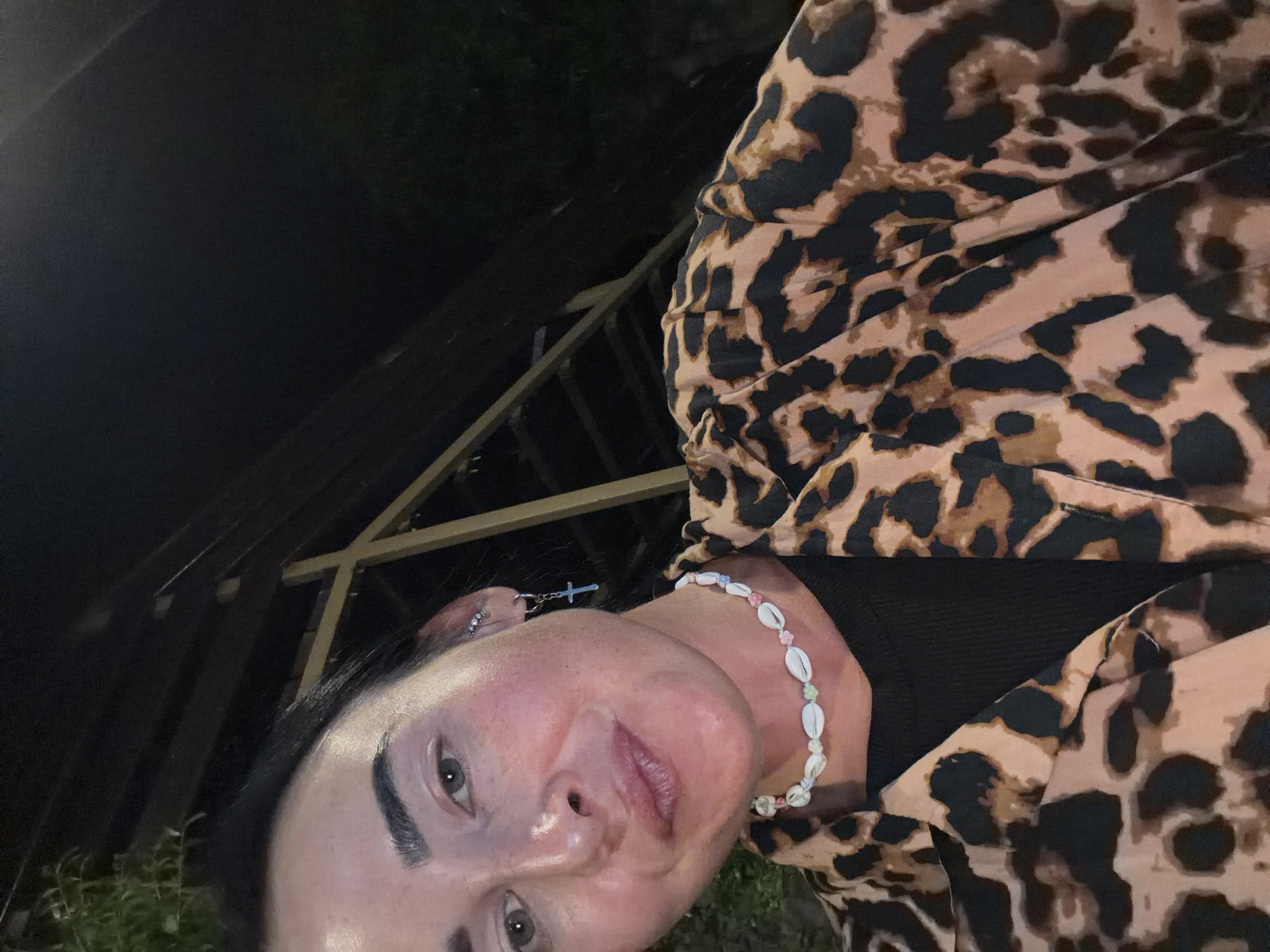 A medium shot shows a woman with dark hair and light-colored eyes posing for a selfie at night, outdoors. She is wearing a leopard-print shirt over a black top, a necklace made of shells and beads, and earrings with a cross pendant. The background is dark and out of focus, with some structure visible, possibly a pergola or outdoor seating area. The scene appears to be in a warm climate, potentially the city of Sochi, Russia, given the presence of tropical foliage and the casual outdoor setting.