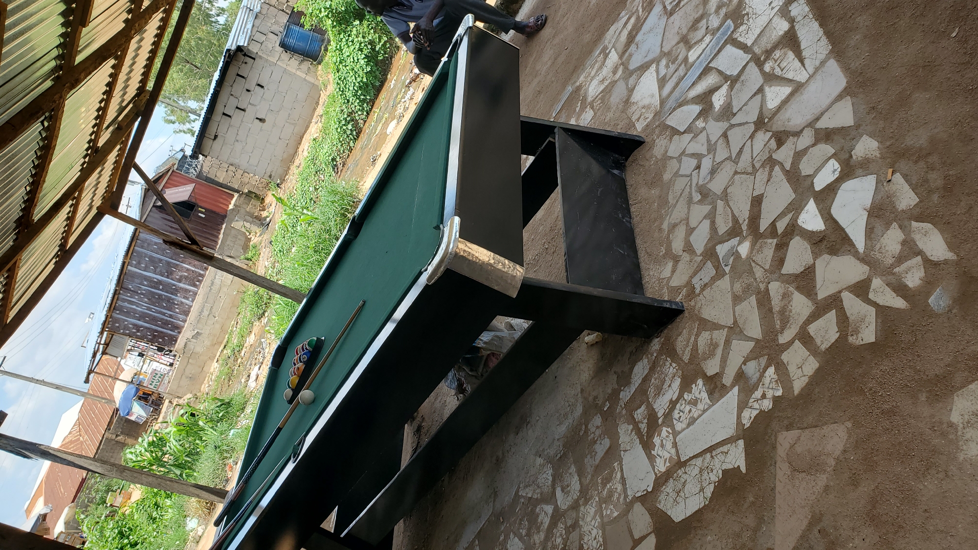 The image depicts a billiard table situated outdoors under a corrugated metal roof. The table is green with a set of billiard balls and two cues arranged on its surface. To the right of the table, a person wearing a grey shirt and dark pants is seated and looking down. The ground around the table is a mixture of sand and broken white tiles. In the background, there are various structures, including buildings with corrugated metal and brick walls, dense green vegetation, and a blue barrel. The overall impression is that of an informal outdoor recreational space, possibly in a less developed area. The presence of residential structures and vegetation suggests an urban or peri-urban environment in Jalingo, Nigeria.