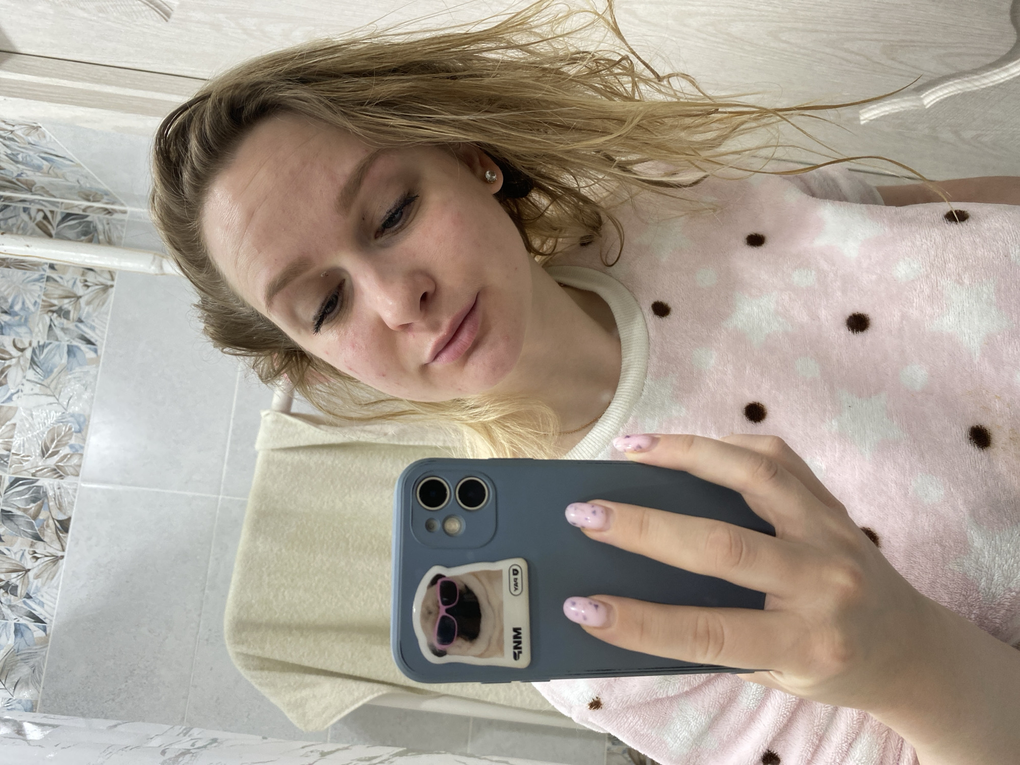 A young blonde woman is depicted in a selfie taken in Chaikovsky, Russia, likely within a bathroom or a room with tiled walls. She is looking downwards towards the smartphone she holds, with a neutral and slightly contemplative expression. Her light-colored hair is somewhat windswept or disheveled around her face and shoulders. Her skin is fair, with some visible redness or blemishes on her cheeks and forehead. She appears to be wearing minimal makeup, with visible eyeliner. A single stud earring is in her right ear.
She is dressed in a soft, fluffy, long-sleeved top or sweatshirt, which is pink with a pattern of white stars and scattered brown polka dots. Her fingernails are manicured with a light pink polish adorned with small darker flecks.
The smartphone she is holding features a blue or grey case with a prominent sticker on its back. The sticker displays a pug dog wearing pink sunglasses. Vertical text "SNM" is visible on the left side of the sticker, and the letters "YAR" are visible but reversed on the top right, indicating a mirror reflection. The phone itself has a dual-lens camera system.
The background shows tiled walls. The upper part of the background features white, horizontally oriented tiles. The lower left portion displays grey tiles interspersed with decorative tiles featuring a blue and brown leaf pattern. A light-colored, possibly off-white, towel or fabric is draped in the mid-ground. The lighting suggests it is daytime.