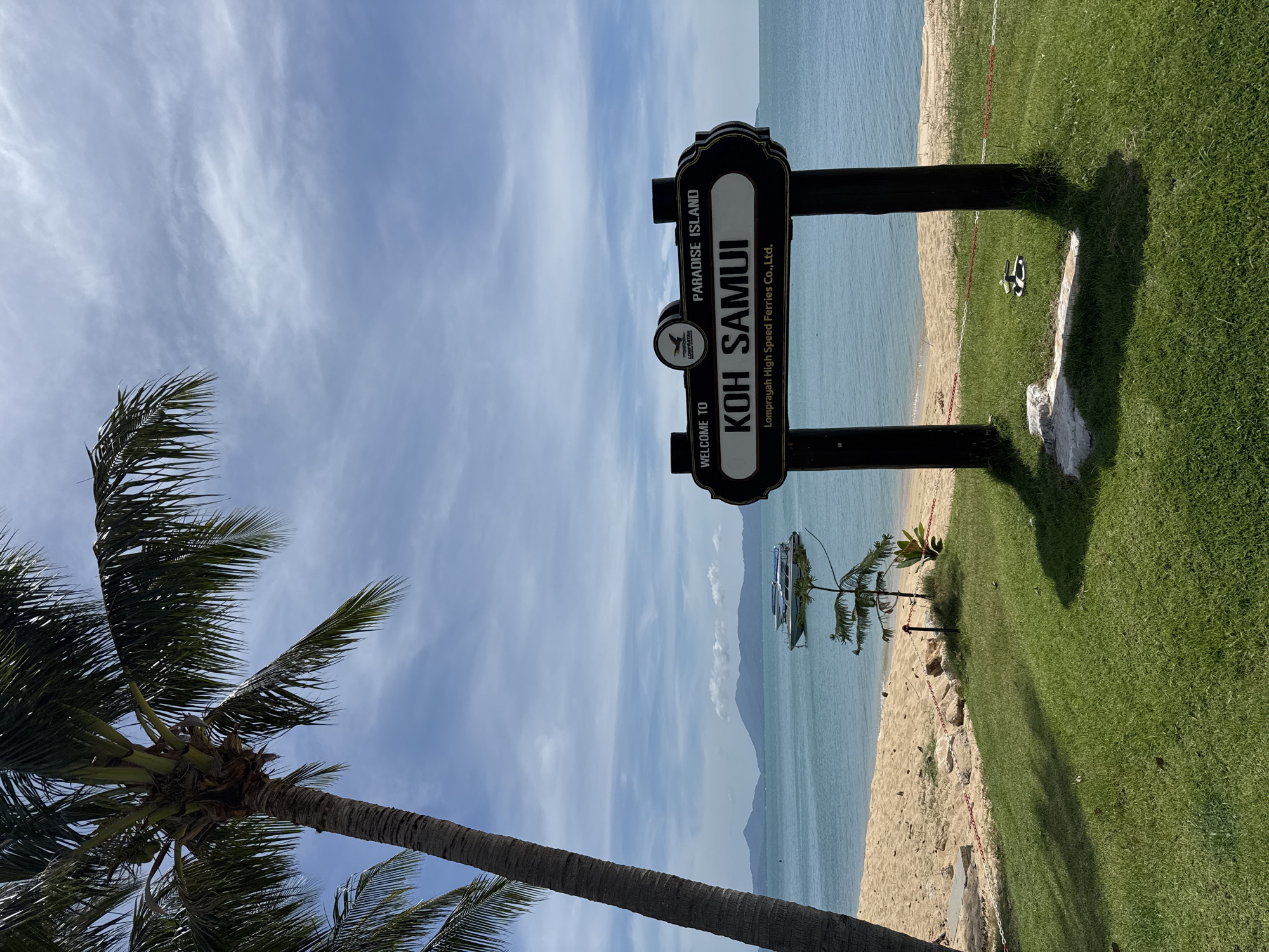 Here is a description of the image:
The image features a sign that reads "Welcome to Koh Samui, Paradise Island," with the words "Lomprayah High Speed Ferries Co., Ltd." also present. This indicates a tourism-related welcoming sign, potentially marking an entry point to Koh Samui, which is an island in Thailand. The location is suggested to be Baan Tai in Thailand.
In the background, there's a view of the beach and the sea under a sky with scattered clouds. A palm tree is visible on the left side of the image, contributing to the tropical ambiance. The foreground includes a grassy area where the sign stands. The sign is supported by a wooden structure. There are boats on the water in the distance, off the beach. A pair of sandals is resting on the grass next to the sign.