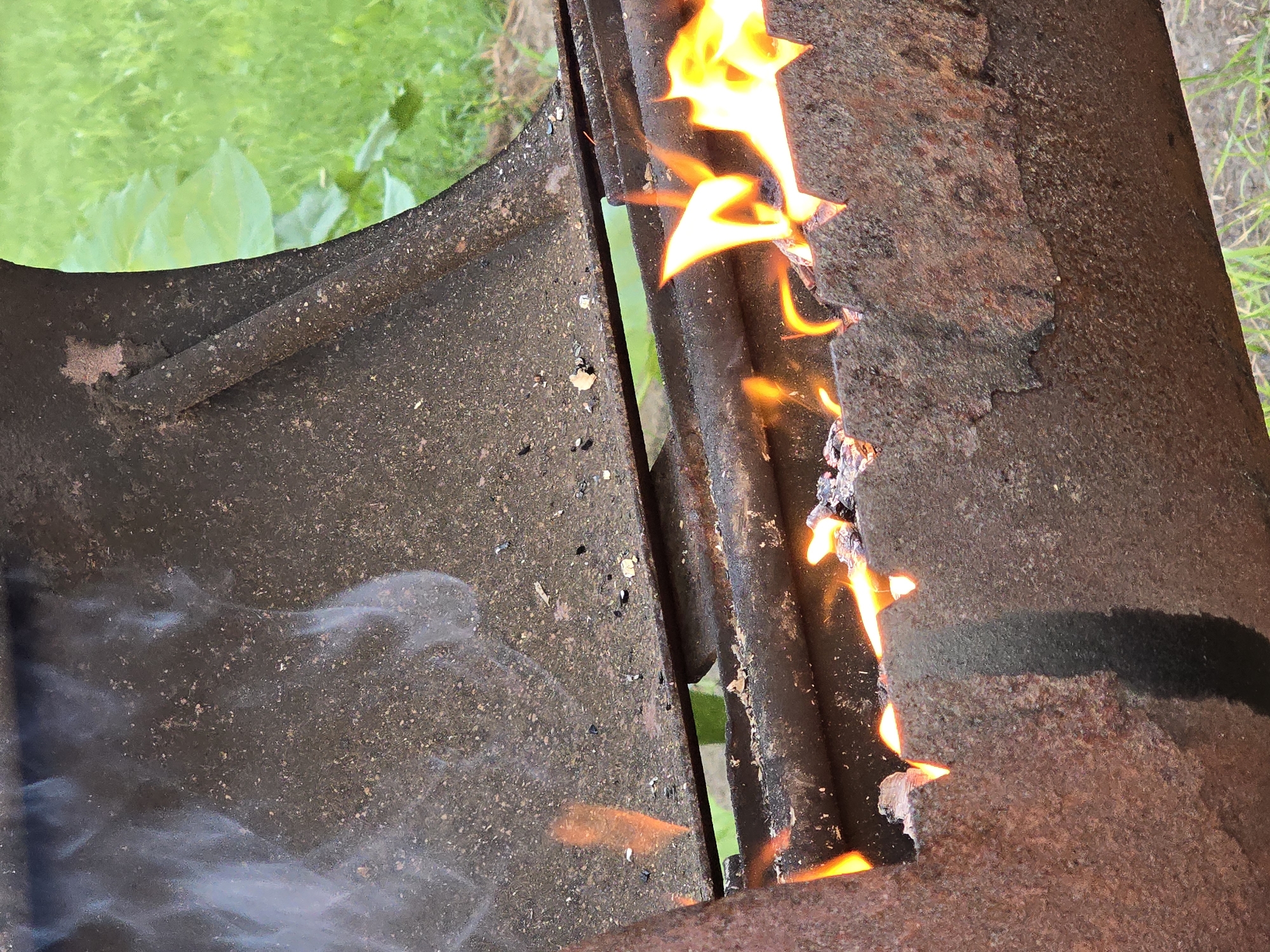 A close-up, waist-level shot shows a rusted metal barbecue grill with an active fire. The grill has a weathered, dark brown exterior with visible patches of rust and pitting. Flames are visible through a broken section of the grill's lower edge, indicating a fire within. Wisps of white smoke rise from the left side of the grill. In the background, blurred green grass and foliage suggest an outdoor setting, possibly a yard. The scene is devoid of any human presence. The location is noted as being in the city of Ермолаево, Russia.