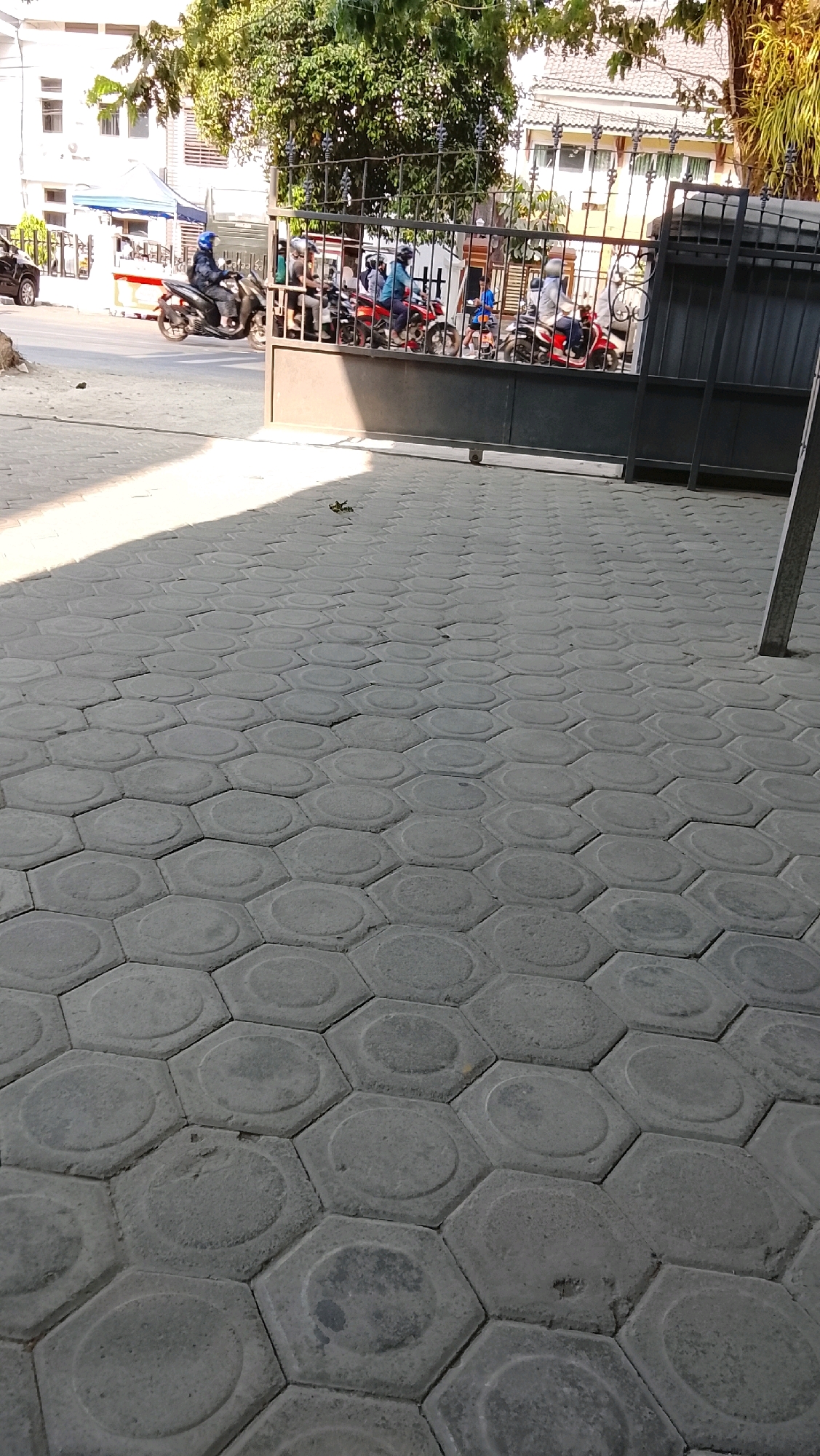 The image shows a street scene in Slawi, Indonesia, with a focus on a hexagonal-patterned pavement in the foreground. In the background, a metal gate is open, revealing several people on motorcycles waiting or perhaps in a line. Trees and buildings are visible behind the gate, suggesting an urban or semi-urban environment. The lighting indicates it is daytime, with sunlight casting shadows on the pavement. A black car is partially visible on the left side of the frame.