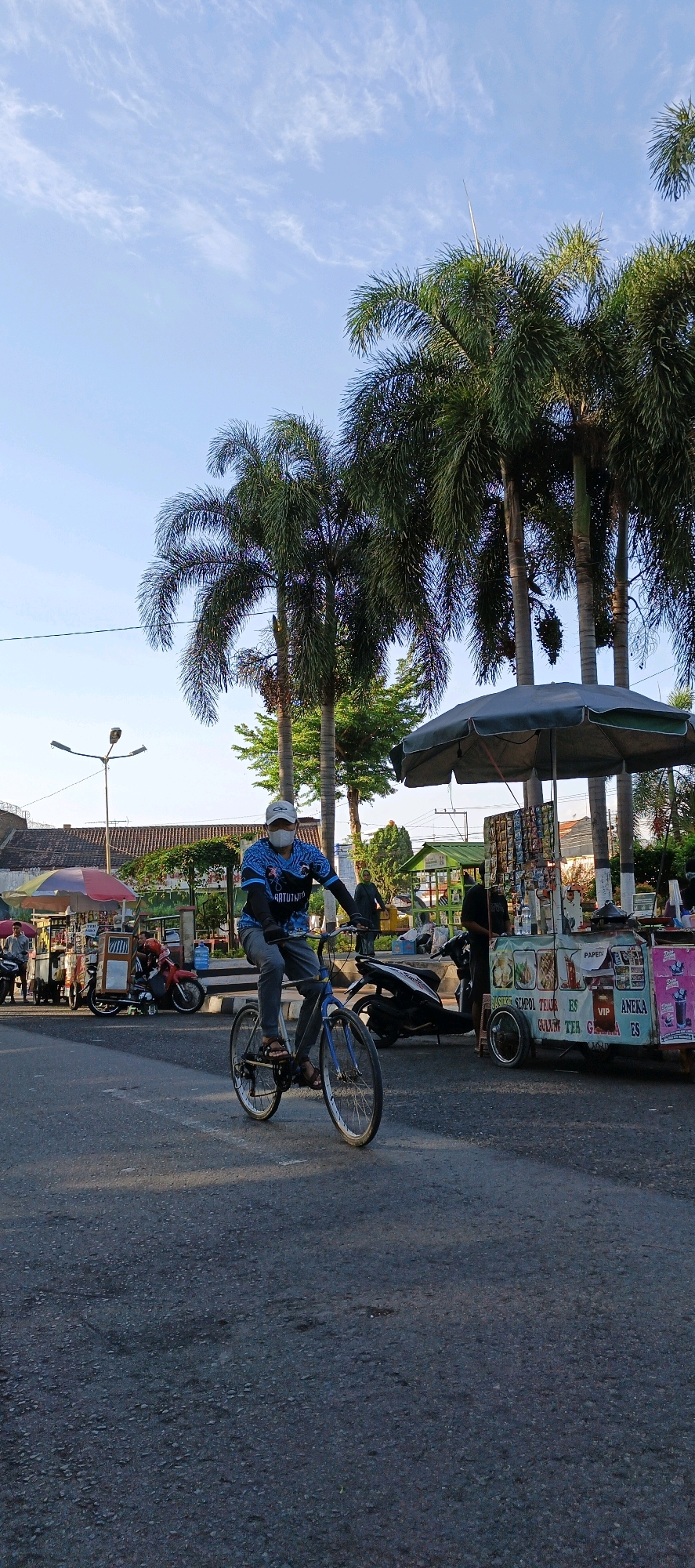 A man wearing a blue shirt, grey pants, a face mask, and a white cap rides a blue bicycle on an asphalt road. He is positioned in the center of the frame, facing slightly to the right. Behind him, a street food cart with an umbrella is visible, stocked with various drinks and snacks. Several palm trees line the background, with a clear blue sky above. To the left of the frame, another food cart and a motorcycle are present, with a street light visible in the distance. The setting appears to be a street in Klaten, Indonesia, likely during daytime with natural lighting.