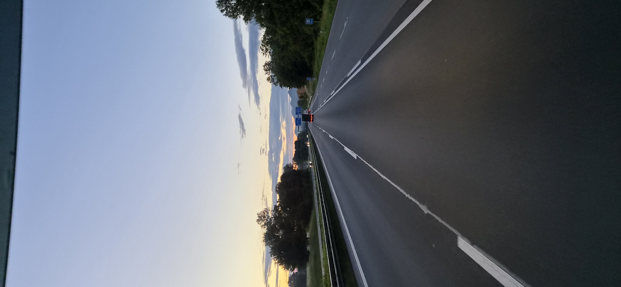 This vertical-format image captures a view from inside a vehicle traveling on a multi-lane highway near Raubling, Germany, during either dawn or dusk. The sky transitions from a light blue overhead to a band of warm yellow, orange, and faint pink hues at the horizon, with scattered grey clouds in the lower sky.
The asphalt highway features at least two lanes in the foreground, marked by white solid and dashed lines. A metal guardrail separates the lanes of travel from an opposing set of two lanes, which also feature a guardrail. In the same direction of travel, a red passenger car is visible in the left lane, and a dark-colored commercial truck is further ahead in the right lane, both displaying illuminated taillights. In the opposite direction, several vehicles are visible in the distance, indicated by their illuminated headlights.
Dense green trees and vegetation line both sides of the highway. On the right, several blue rectangular road signs are visible in the middle distance, along with a smaller blue sign closer to the foreground. Beyond the trees and signs on the right, distinct mountains or large hills are silhouetted against the colorful horizon. The very top edge of the image is obstructed by a dark, horizontal element, likely part of the vehicle's interior. No people are discernible.