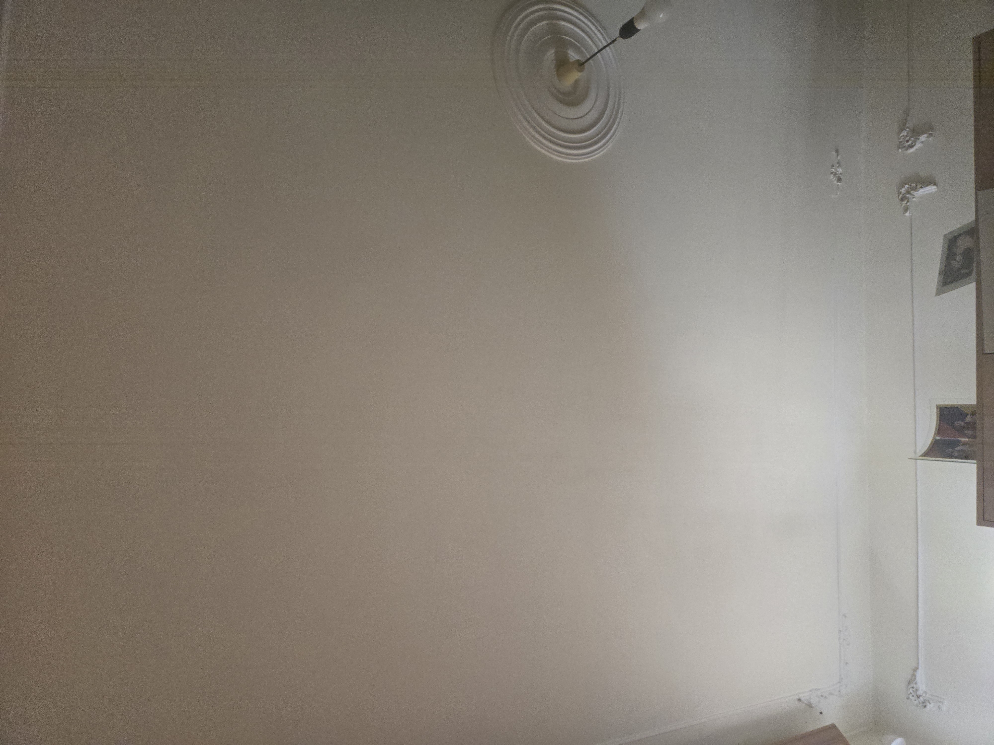 This media file presents an interior view of a room, likely in a residential setting in Yerevan, Armenia. The central subject is a light-colored, plain wall, possibly painted off-white or beige, which dominates the left and central parts of the frame.
On the ceiling, a decorative, circular white ceiling rose with concentric rings is prominently featured. A single, bare light bulb hangs from its center by a dark wire, providing illumination to the room.
To the right, a corner of the room is visible, revealing a perpendicular wall. This wall is adorned with several small, ornate white decorative pieces, likely plasterwork or carved wood, resembling floral motifs or abstract patterns, positioned at different heights. Below these, a framed picture, possibly a portrait, is partially visible, leaning against or placed on a vertical wooden structure that appears to be part of a cabinet or shelving unit. Another similar decorative element is located lower on this right wall, near a white baseboard. The overall lighting is soft, suggesting diffused natural light or interior lighting, with the main wall showing a slight gradient from brighter on the right to slightly darker on the left.
No people are present in the image, and no specific activity is taking place. There is no visible text. The room's aesthetic appears functional with traditional decorative accents.