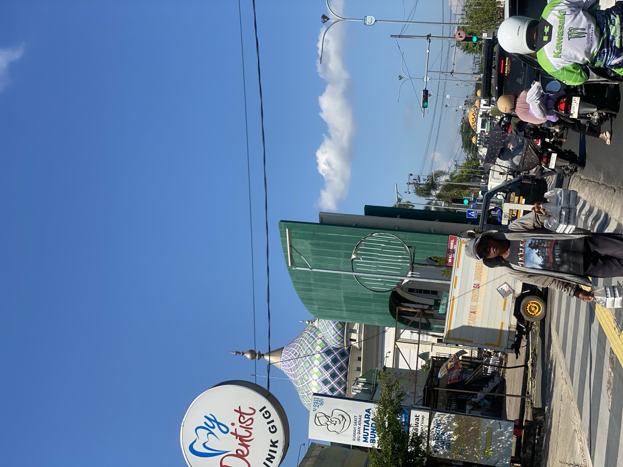 A street scene in Jawa, Indonesia, features a bright blue sky with scattered clouds. In the foreground, a person walks across a crosswalk, carrying two plastic bags of bottled water. They are wearing a gray hooded jacket over a graphic t-shirt and dark pants. Behind them, traffic moves along the road, including motorcycles, cars, and a white truck with "PT. ARTIKA SENTOSA INDONESIA" printed on its side. To the left, a sign for "My Dentist" and a billboard for "RUMAH SAKIT IBU DAN ANAK MUTIARA BUNDA" are visible. Further in the background, the distinctive domes of a mosque, adorned with green and white geometric patterns, punctuate the skyline. Streetlights and power lines crisscross the upper portion of the image.