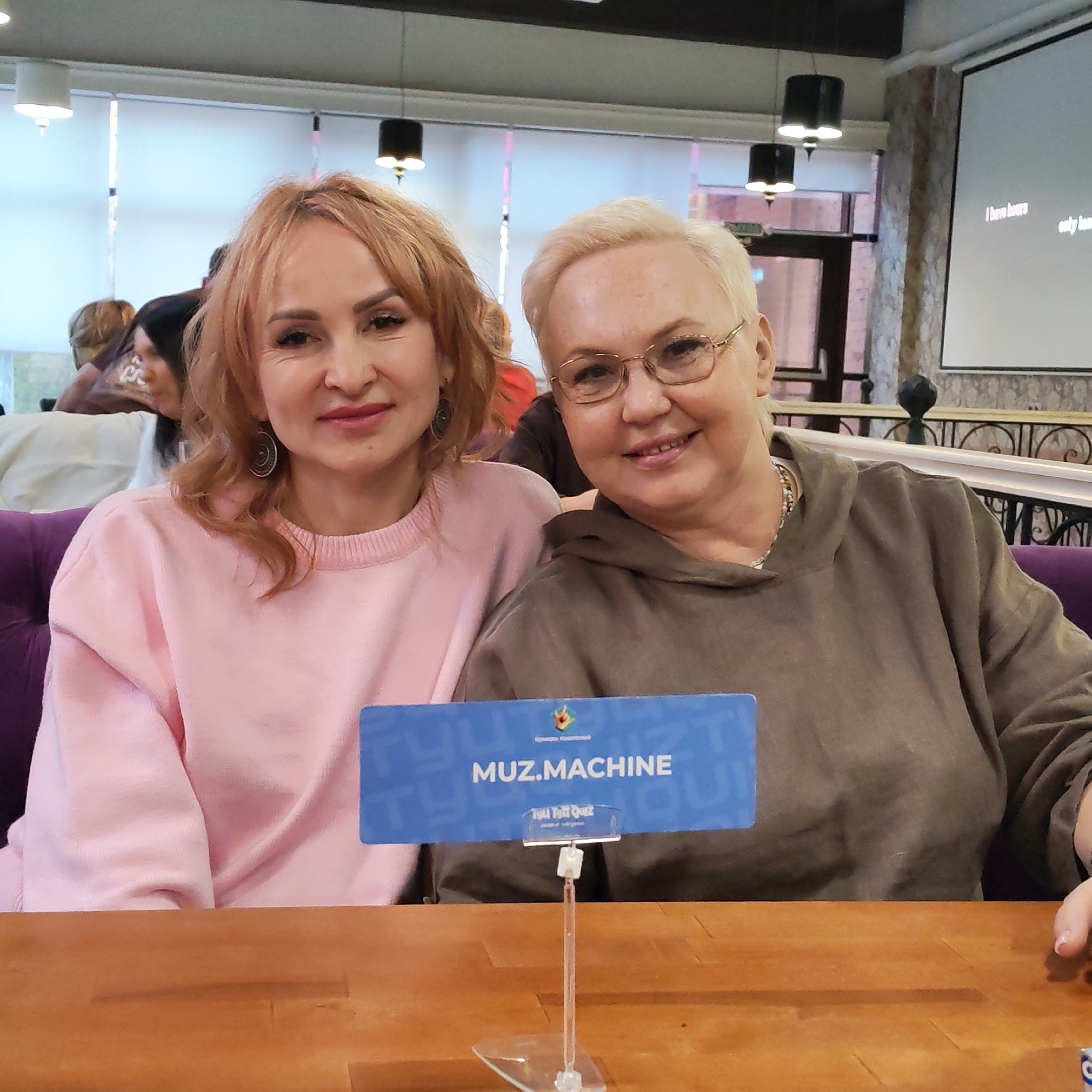 Two women are seated at a wooden table in what appears to be a restaurant or cafe. The woman on the left has reddish-blonde hair and wears a light pink sweatshirt and round earrings. The woman on the right has short blonde hair, glasses, and is wearing an olive green hoodie. In front of them on the table is a sign that reads "MUZ.MACHINE" in blue lettering. They are seated on a purple velvet bench. In the background, other patrons are visible, along with lighting fixtures, a projector screen displaying words, and architectural details such as metal railings. The location is Chaikovsky, Russia.