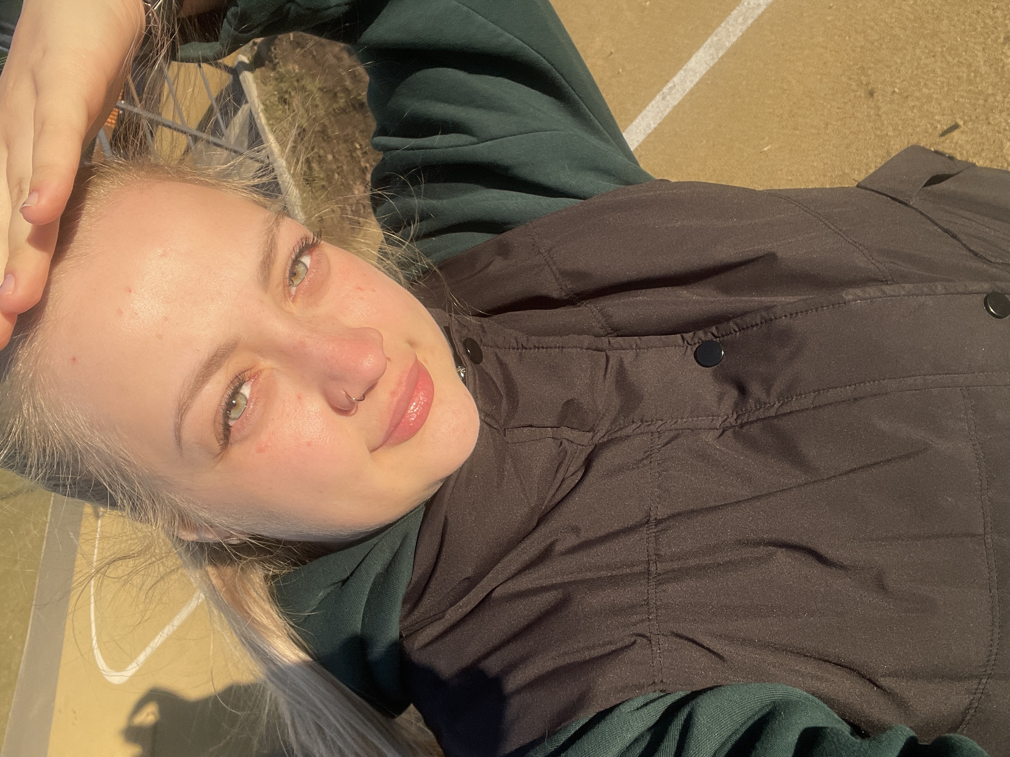 A young woman is lying on a paved surface marked with white lines, possibly a sports court, in Izhevsk, Russia. She is wearing a dark green hooded sweatshirt underneath a black quilted vest. Her blonde hair is partially obscuring the view, and her hand is touching her forehead. She has fair skin and green eyes, and a nose ring is visible. The scene is bathed in sunlight.