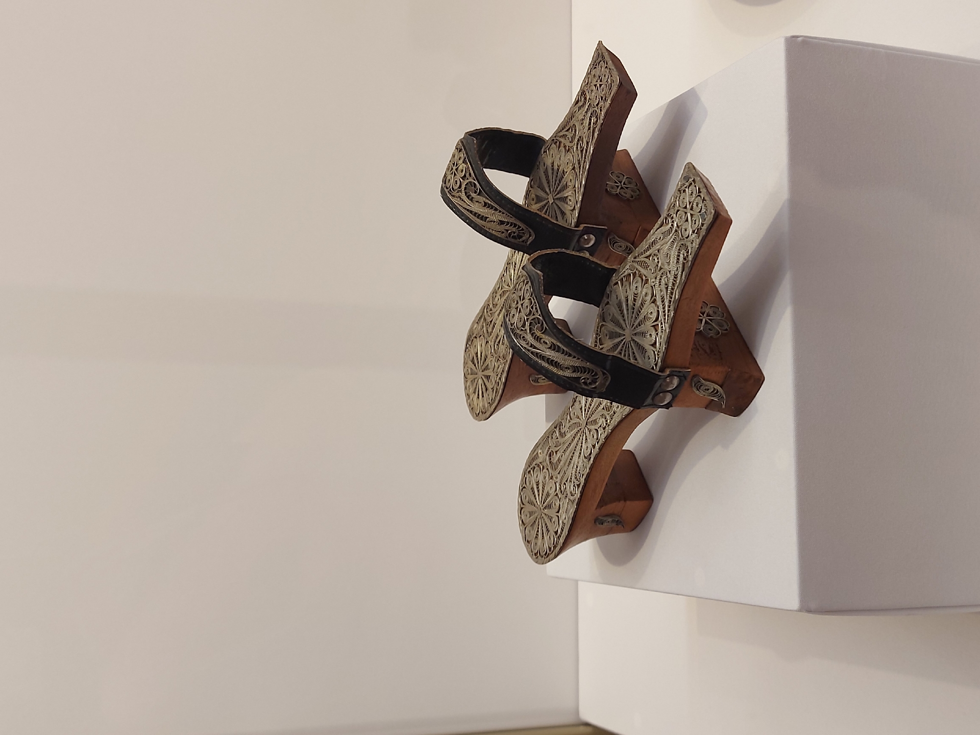 Two wooden shoes with decorative silver filigree and black straps are displayed on a white platform against a light-colored wall. The shoes are positioned one behind the other, showcasing the intricate designs. The scene appears to be an exhibit in a museum or gallery, although the specific location within Saint Petersburg, Russia is not discernable from the image.