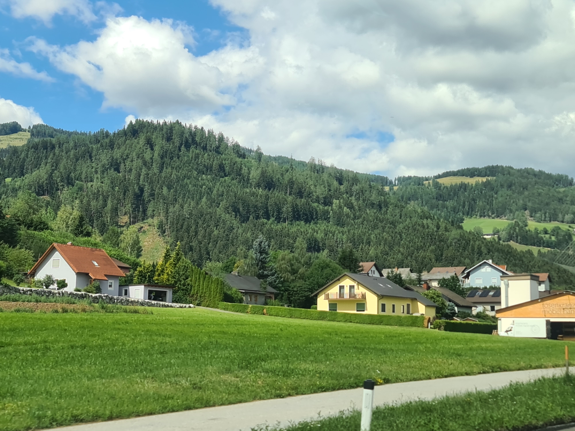The image depicts a rural scene in the vicinity of St. Peter ob Judenburg, Austria. A gently sloping, well-maintained green lawn occupies the foreground. A paved road or path runs along the bottom edge of the frame. In the mid-ground, several houses are visible. To the left, a light grey house with a red-tiled roof and a detached garage is situated on a slight incline, bordered by a stone wall and well-kept hedges. Further to the right, a bright yellow house with multiple windows and a balcony stands, also surrounded by manicured greenery. Behind this, a collection of other residences with various rooflines and colors are nestled amongst trees. A large, densely forested hill or mountain forms the backdrop, stretching across the upper half of the image. The sky above is blue with scattered white, fluffy clouds, suggesting fair weather. The overall impression is one of a peaceful, residential area integrated into a natural, hilly landscape.
