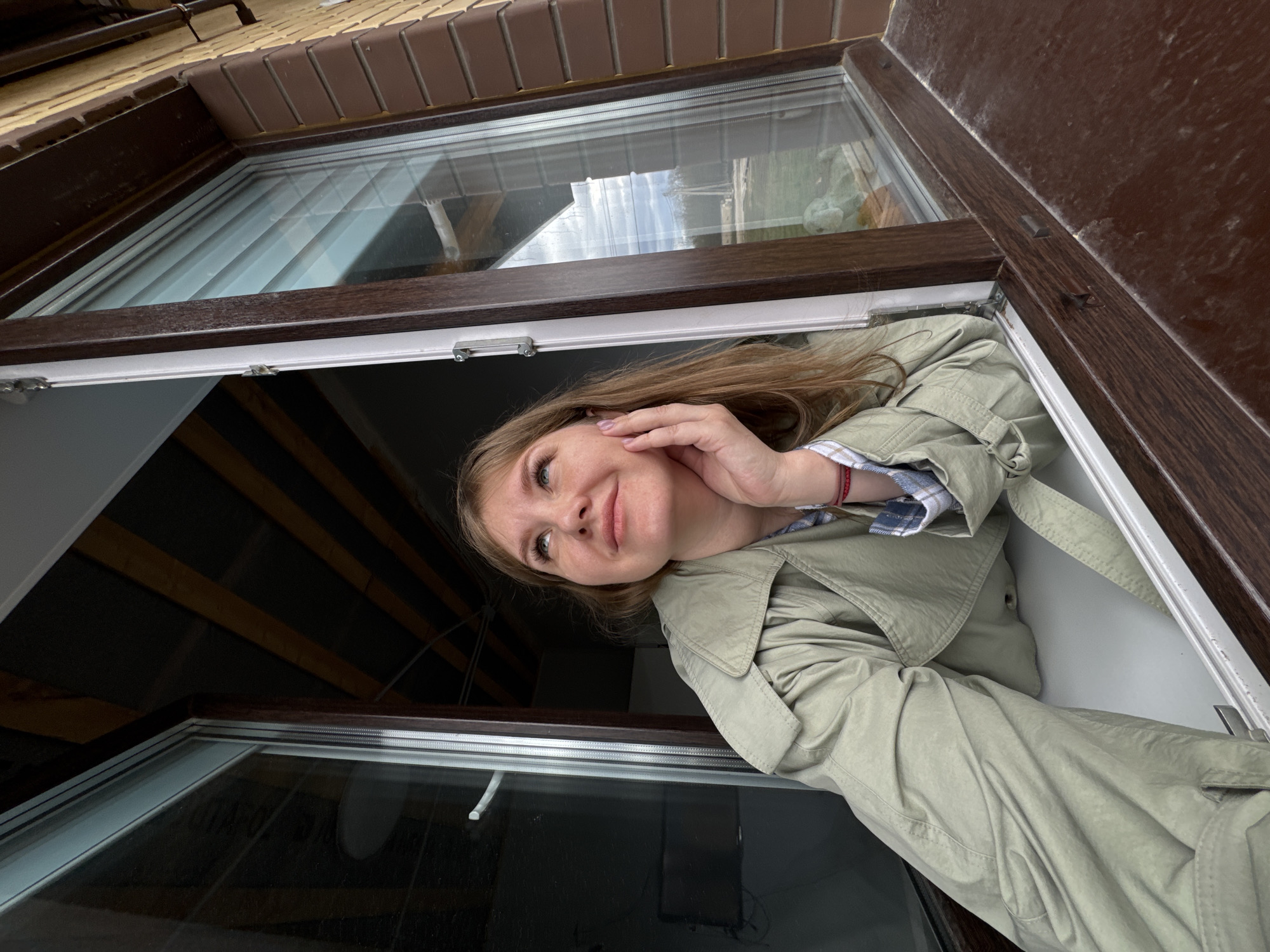 A young woman is captured in a close-up shot, leaning out of a window at an unusual, tilted angle, giving the impression that she is looking upward towards the camera. Her head and upper torso are visible, framed by the window's opening. She has light brown hair, fair skin, and light-colored eyes, and wears a light olive or beige trench coat over a plaid shirt. Her right hand is gently placed against her cheek, and she has a soft, pleasant smile on her face. A red bracelet is visible on her left wrist.
The window has a dark brown, possibly wooden or wood-effect, frame with a white inner trim. The glass reflects parts of an overcast sky and possibly interior structures. Outside the window, a light brown brick wall is visible above, with a darker brown trim or eaves board. Inside, dark wooden beams or rafters are visible in the background, suggesting an attic or upper floor setting. The window itself appears to be a skylight or roof window, opening outwards or upwards.
The scene takes place during the daytime, indicated by the natural light and cloud reflections in the windowpane. The weather appears to be overcast or partly cloudy. The overall mood conveyed by the woman's expression is one of calm and contentment. There is no visible text in the image. The location context is Gornyi (Dvurechenskoe S/P), Russia.