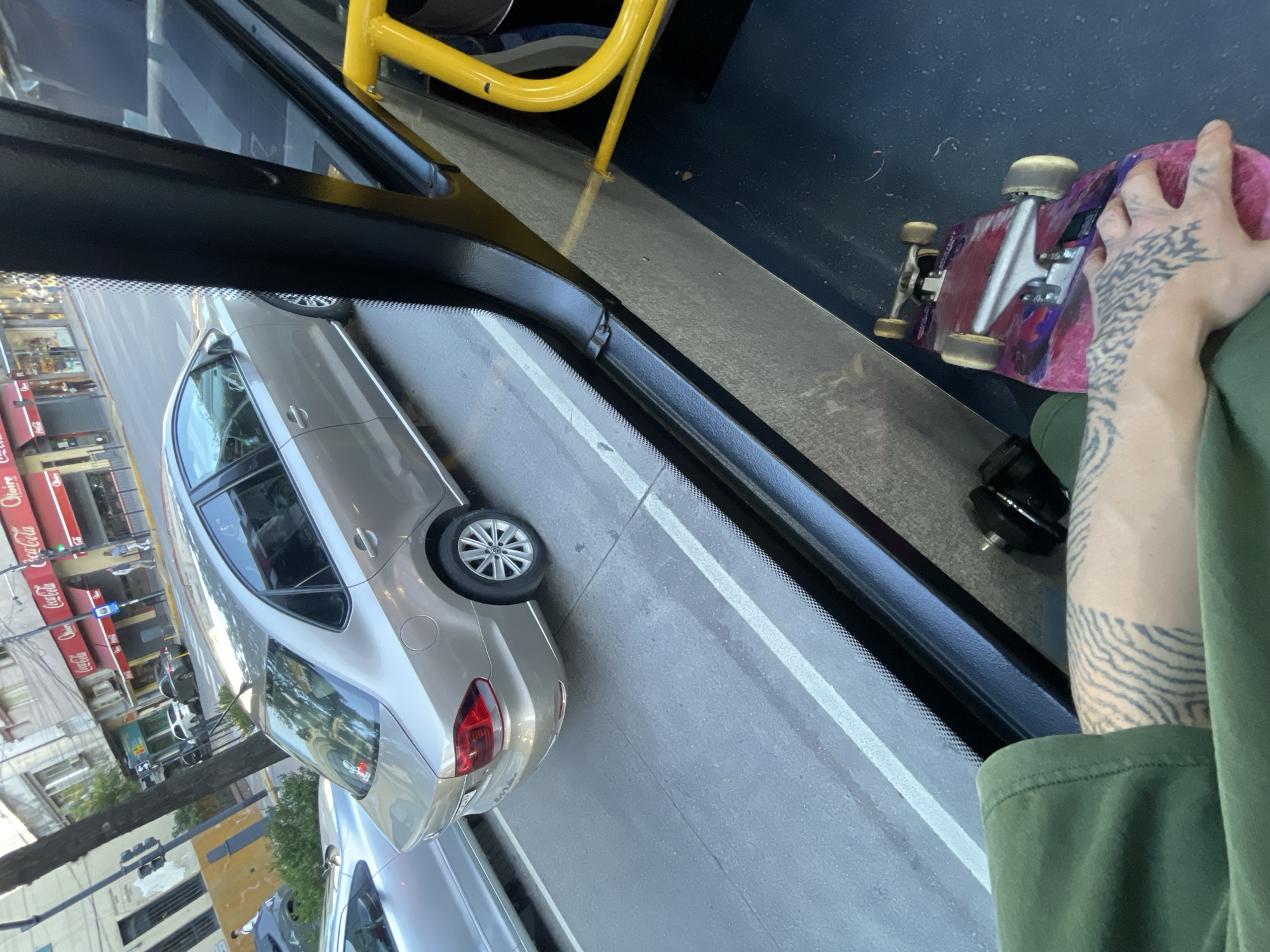 From the perspective of a person on a bus, a view of Vicente López, Argentina is observed. A gray car is visible outside the window. The person on the bus has a skateboard and a tattoo on their arm. Coca-Cola signage is visible on a building.