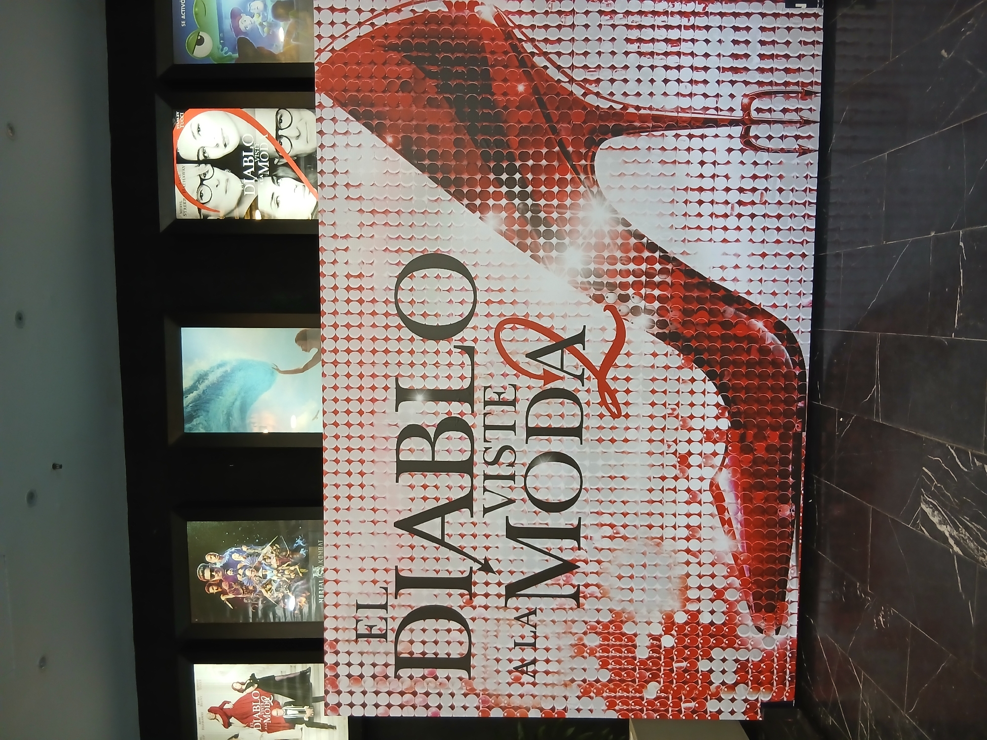 The image shows a movie poster display in an indoor setting, possibly a cinema or entertainment complex. The posters are arranged vertically on a black wall. Starting from the top, the posters show: an animated children's film, a poster featuring Meryl Streep and Anne Hathaway that says "El Diablo Viste a la Moda," an image of a woman in water, a poster for "Mortal Kombat," and then another poster for "El Diablo Viste a la Moda." The wall is adjacent to a marble wall. The setting is unclear, but this image does not contain information about the city of Altagracia, Venezuela.