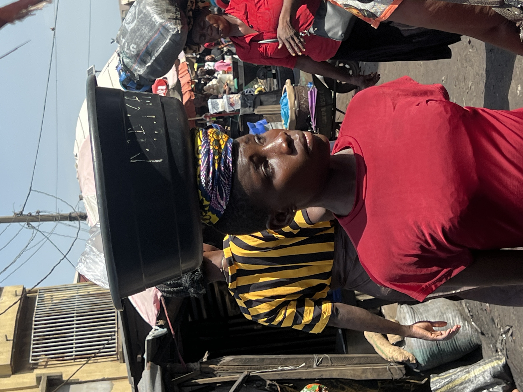 In a street in Nkpor, Nigeria, a woman in a red shirt balances a large black basin on her head, secured by a colorful head wrap. She gazes upwards, while another woman in a striped yellow and black shirt stands beside her. A third woman in a red shirt is visible in the background, along with various goods and belongings, including large bags and other objects. The environment appears to be a bustling market or street scene with buildings visible in the background. Power lines are visible in the sky.
