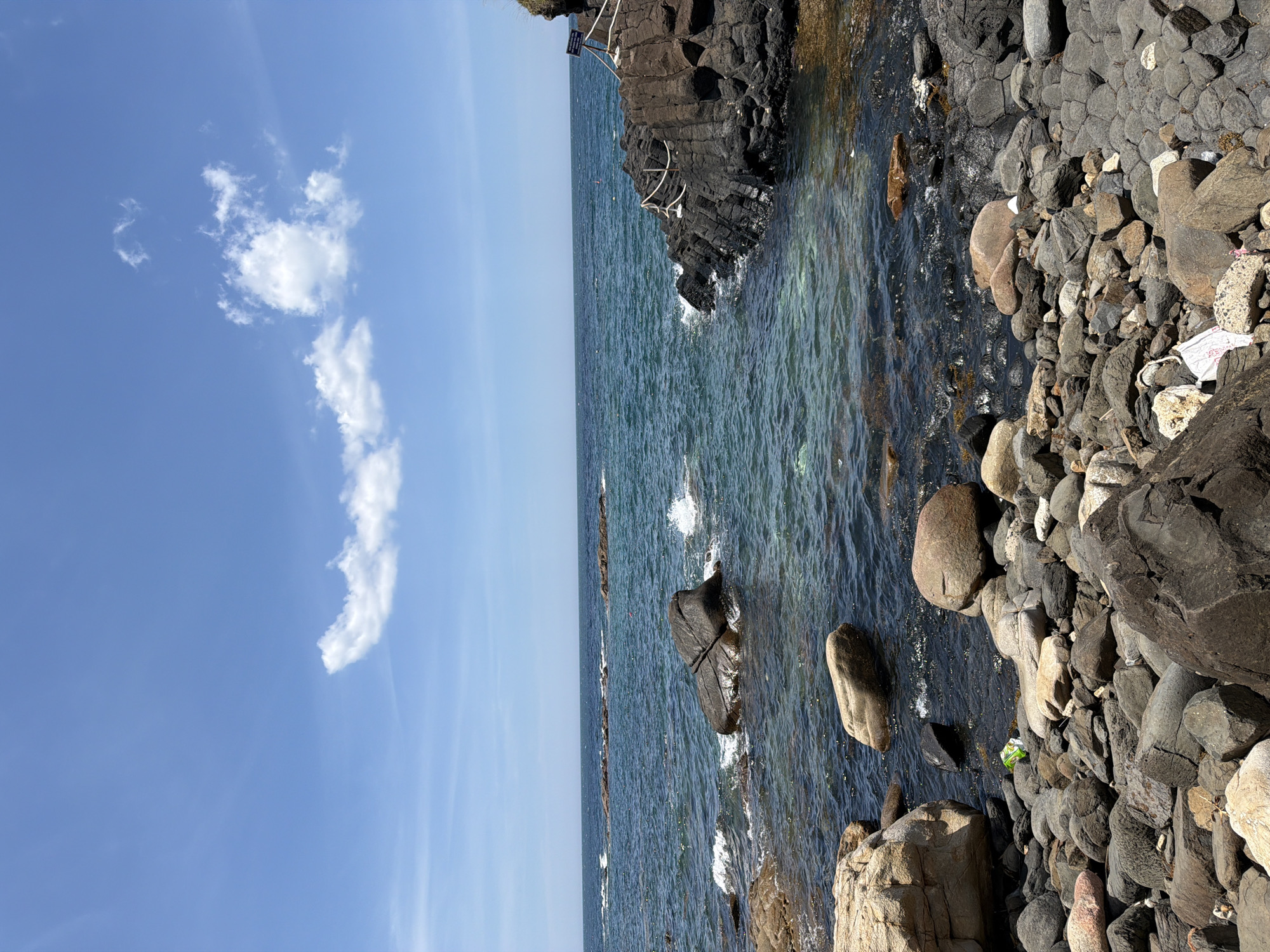 The image depicts a rocky coastline under a clear blue sky with scattered white clouds. The sea is a deep blue-green, with waves gently lapping against the shore. The foreground is dominated by a jumble of rocks of various sizes and colors, some smooth and rounded, others rough and angular. In the water, larger rocks are visible, some breaking the surface and creating small splashes. To the right, a black, columnar rock formation extends into the sea, with a white rope railing suggesting a pathway or viewing area. A sign is mounted on a post near the railing. The environment appears natural and somewhat rugged. There are no people visible in the image. The presence of some white and green litter on the rocks suggests human activity, despite the absence of people at the moment. The scene is a snapshot of a coastal landscape, likely a tourist spot given the railing and sign. The location is identified as Phú Lương, Vietnam.