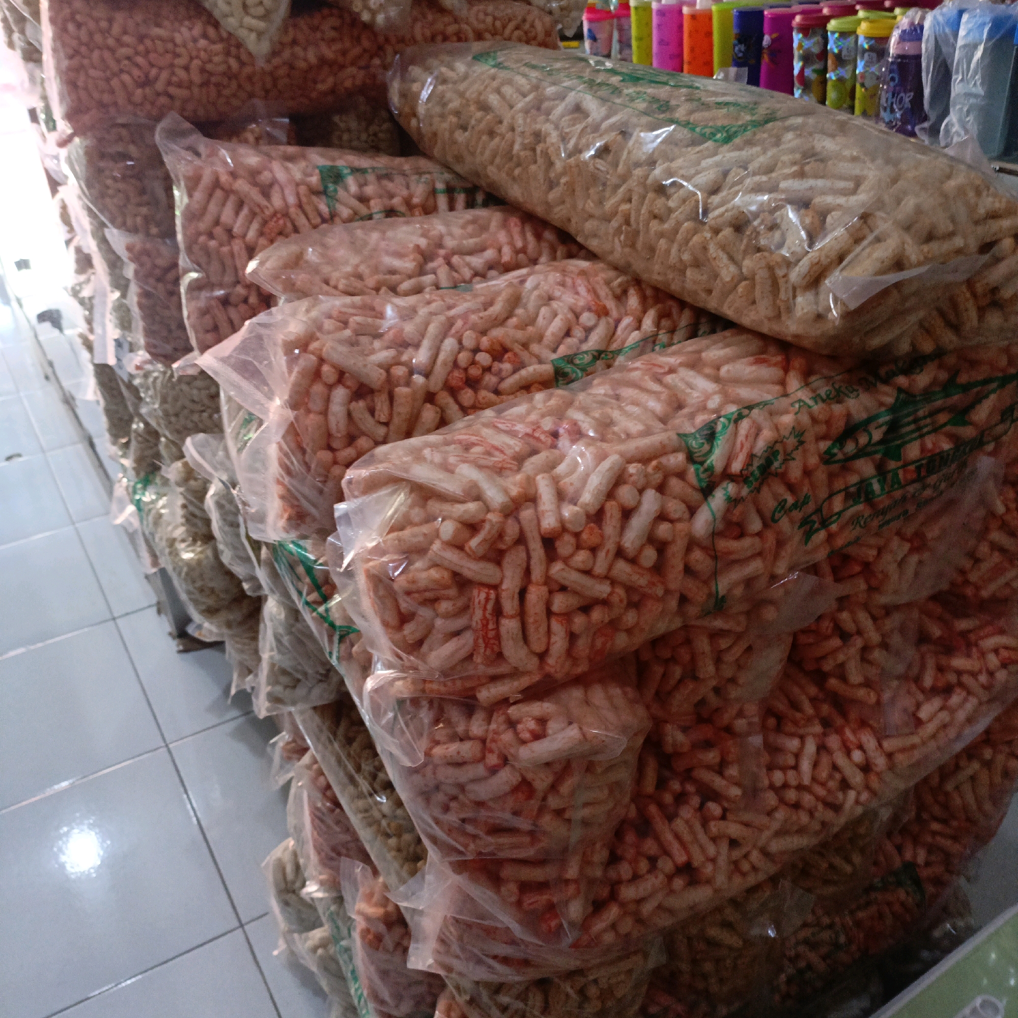 In a retail setting, likely a small shop or market stall, a variety of snack foods are displayed in clear plastic packaging. The snacks, appearing to be extruded and possibly fried, are stacked in large bags. Some bags contain snacks that are light brown or beige in color, while others are a lighter pinkish-orange hue, indicating potential flavoring or seasoning. A few of the bags have green print, possibly branding or product information, on them. In the background, out of focus, are rows of colorful bottles, suggesting they may contain beverages or other retail items. The floor beneath the display is tiled in a light grey color with visible grout lines. The overall scene captures a typical point-of-sale environment for packaged snacks. This scene could be located in Salatiga, Indonesia, a city known for its local food vendors and markets.