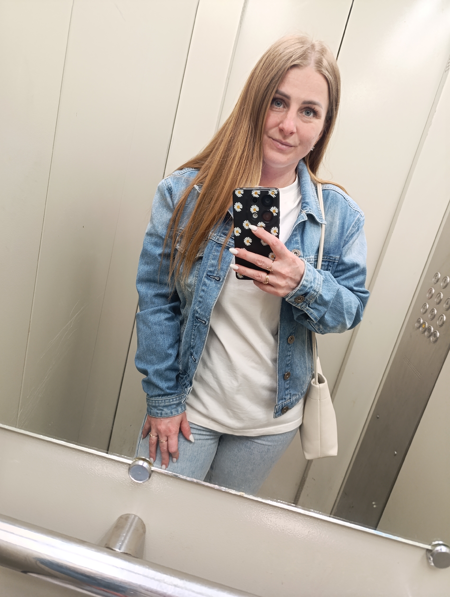A woman is taking a selfie in what appears to be an elevator. She has long, light brown hair, blue eyes, and is wearing a light blue denim jacket over a white t-shirt, light blue jeans, and has a small white purse slung over her shoulder. She is holding a black smartphone with a white daisy pattern case. The elevator has a mirror in which the woman is taking the selfie, a control panel with circular buttons, and a metal handrail near the bottom of the mirror. The location is Unknown, Unknown.