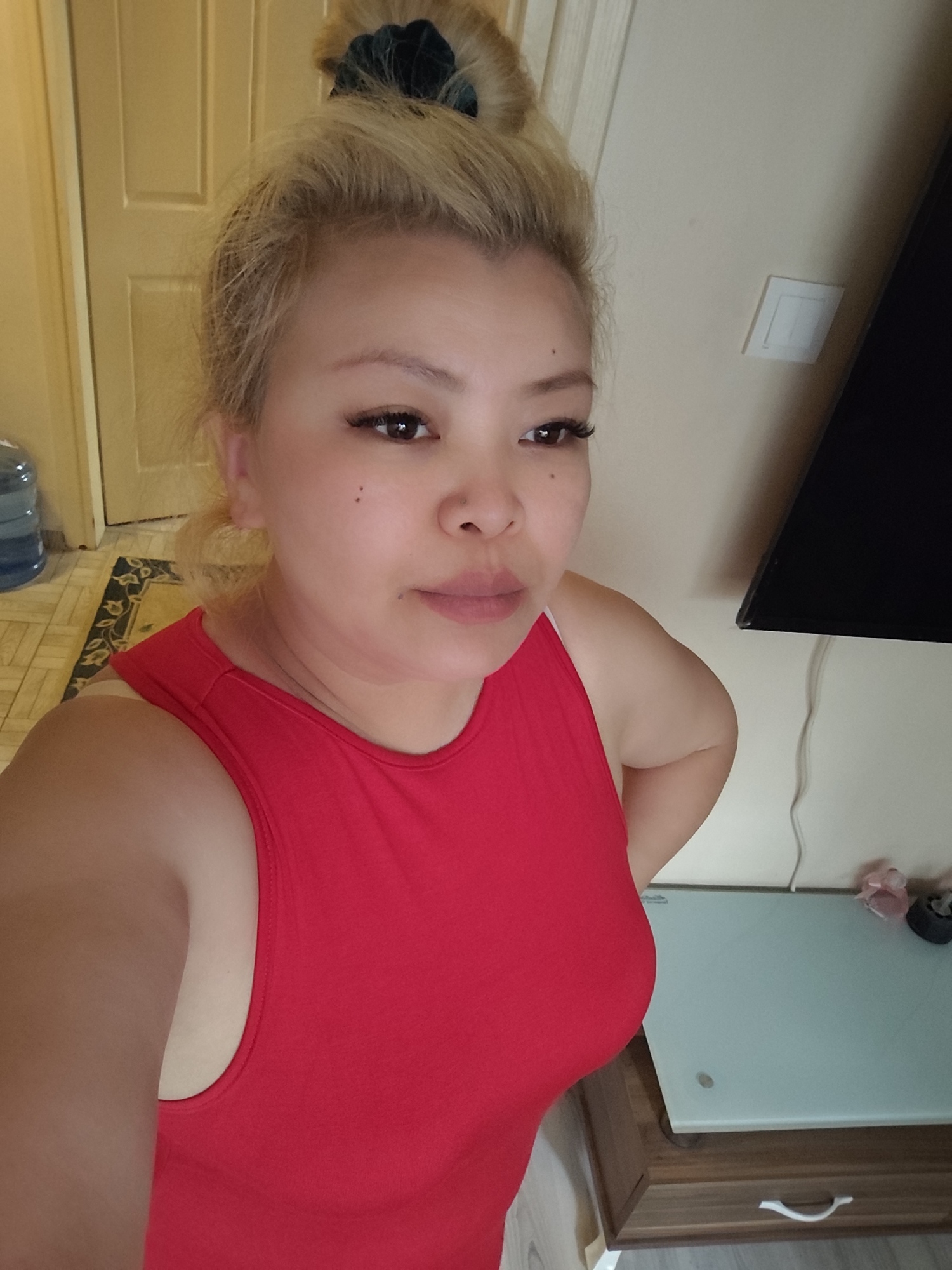 A woman with blonde hair in a bun is posing in a room. She is wearing a red sleeveless top. Behind her is a door, a TV, a light switch, a table, and part of a rug. The location in the city of Sarıyer, Turkey is not discernible from the image.
