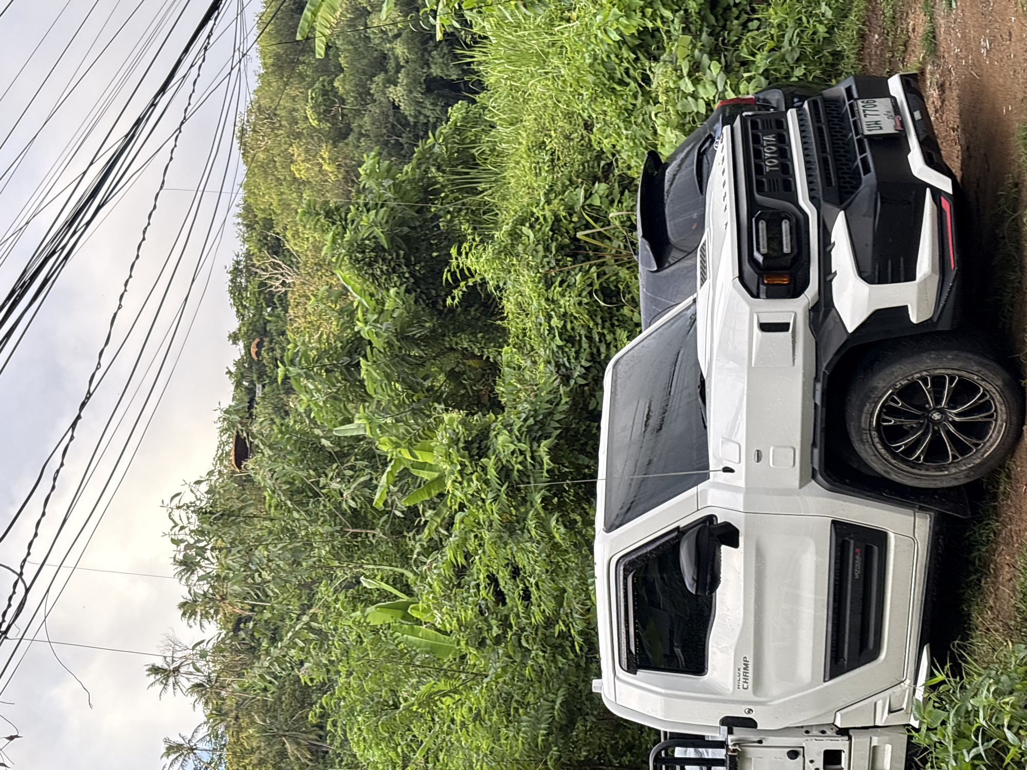 In Karon, Thailand, a white Toyota Hilux Champ truck is parked on a dirt road. The truck features a rugged design with a black grille and accents. A black hatchback is parked behind it. Lush green foliage and trees cover a hillside in the background, with power lines crisscrossing the sky. The scene appears to be outdoors, possibly on the outskirts of the city, given the natural environment.