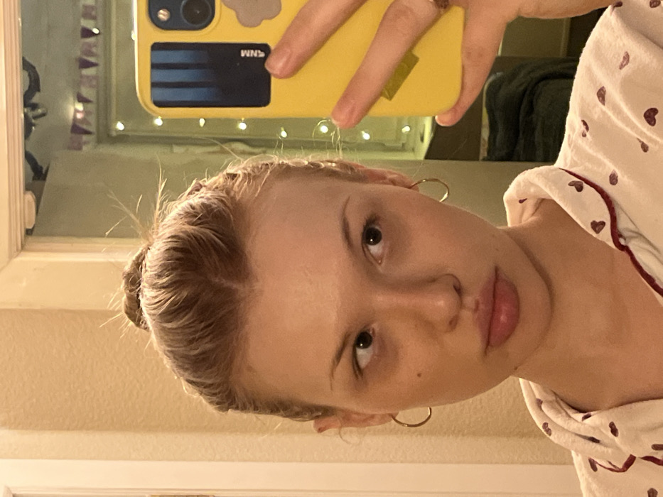 A fair-skinned female subject is depicted in a mirror selfie, holding a yellow-cased smartphone with her right hand. Her light brown hair is pulled back into a messy bun. She wears small, gold-colored hoop earrings and light-colored pajamas adorned with a red heart pattern and red piping. Her face appears without makeup, exhibiting visible dark circles under her eyes and a small mole on her right cheek. A faint line or scar is present on her upper lip, above the cupid's bow. Her dark eyes are directed towards her right, away from the camera lens. The smartphone case features a black rectangular camera module with two lenses, vertical blue stripes on a dark background, inverted "MNM" text, and a light cloud-shaped sticker on the lower right. The background reflection reveals a light-colored wall and a white doorframe to the left. To the upper right, beyond the doorframe, a room is visible, illuminated by a string of fairy lights and decorated with a banner of triangular flags. A dark textile, possibly a blanket, is also reflected. The specific location within Chaikovsky, Russia, cannot be determined from the visual evidence provided.