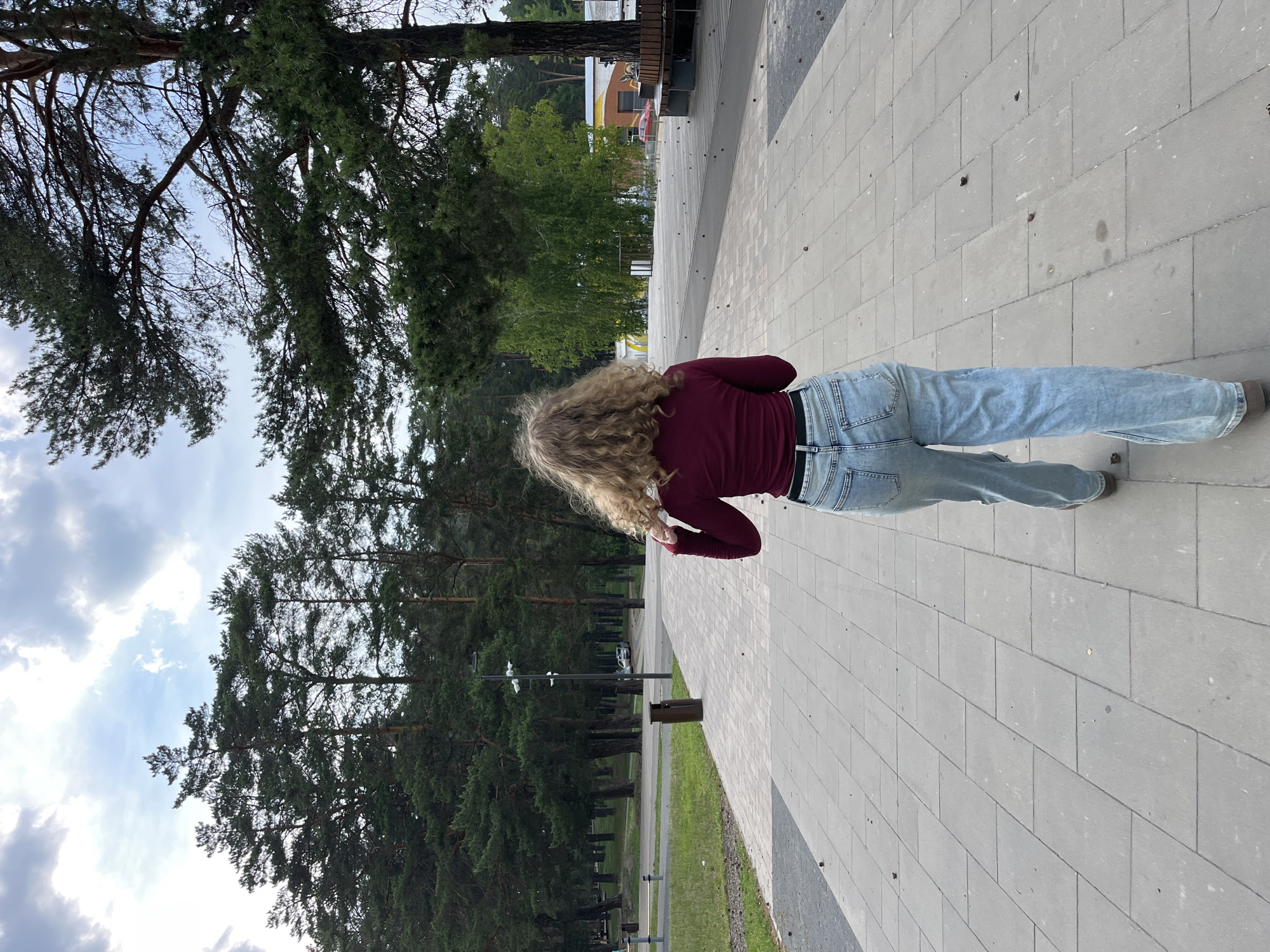 A young woman with curly blonde hair walks away from the camera on a paved walkway in Zelenodol'sk, Russia. She is wearing a maroon long-sleeved shirt and light-wash baggy jeans. The walkway is lined with large pine trees. In the background, there are more trees, a body of water, and some buildings. The sky is cloudy. The overall impression is one of a peaceful, natural setting within a city.