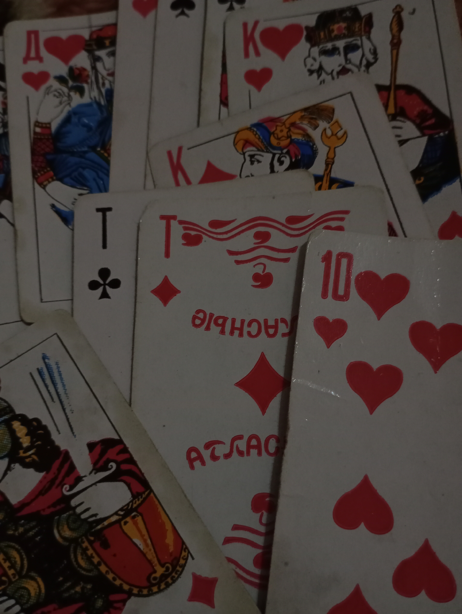The image displays a close-up, top-down view of multiple scattered playing cards, consistent with a Russian "Atlas" pattern deck, featuring Cyrillic letters and distinct court card artwork. The cards are arranged in an overlapping, unorganized fashion on an indistinct, light-colored surface that serves as the background. Prominently visible cards include a Queen of Hearts, marked with the Cyrillic letter "Д" (Dama) and depicting a woman holding a red rose; a King of Hearts, marked with "К" (Korol) and illustrating a bearded man with a scepter; an Ace of Clubs, denoted by "Т" (Tuz) and a club symbol; and a 10 of Hearts, identified by the numeral "10" and multiple heart pips. Another partially obscured card shows a diamond suit and the inverted Cyrillic text "КАСНЫЕ АТЛАС" (Krasnye Atlas). The cards exhibit minor wear along their edges and surfaces, suggesting use. No individuals are present in the scene, nor are specific actions or interactions taking place other than the static arrangement of the cards. The specific location within Kurganinsk, Russia, is not identifiable from the provided visual information.