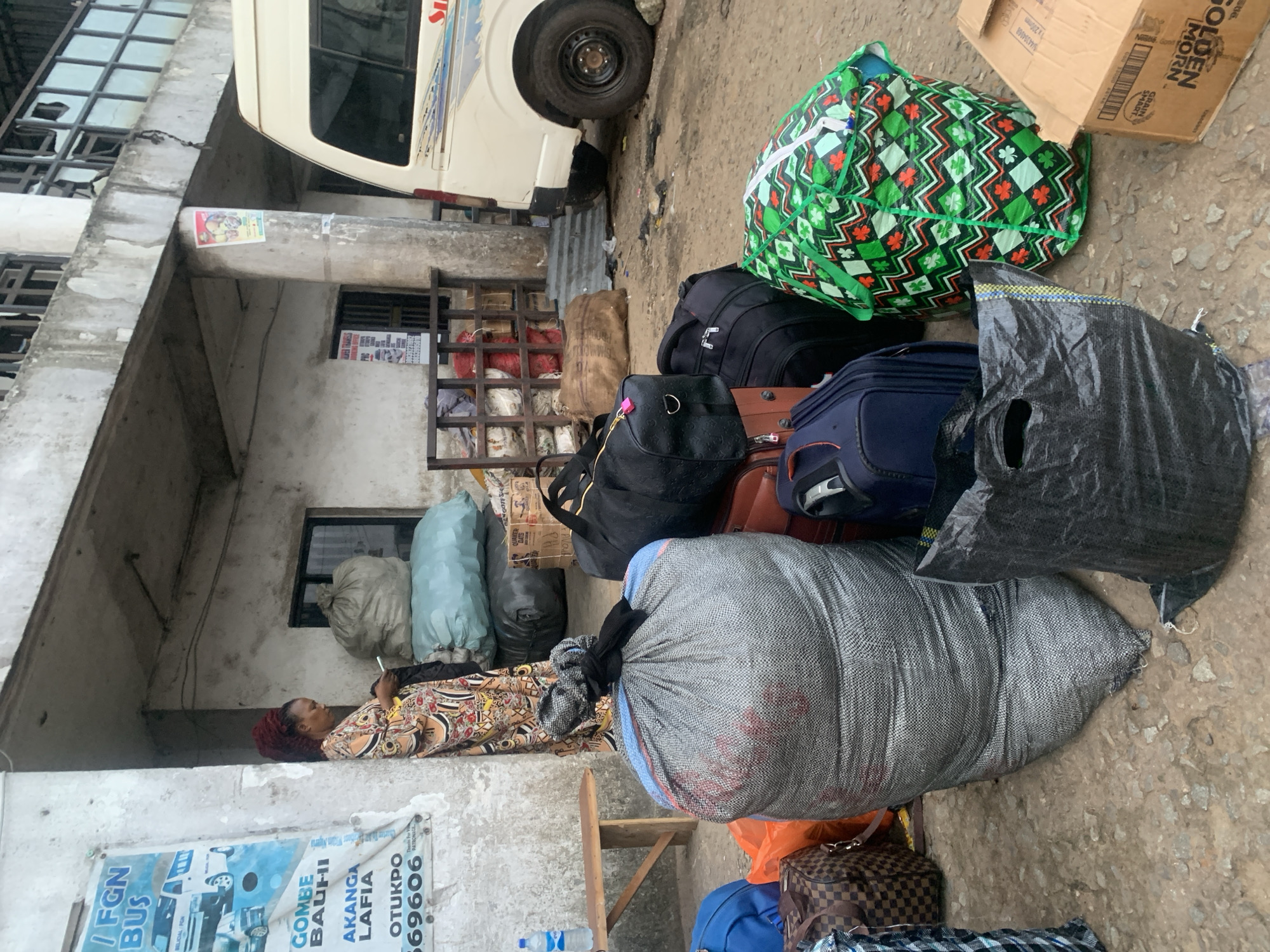 This is a medium shot, eye-level photograph taken outdoors at what appears to be a transportation hub in Elelenwo, Nigeria. The scene is characterized by a mix of luggage and people gathered near a building and a vehicle.
In the foreground, there is a large, grey, woven sack with red markings and a black knot on top. To its right, a large black plastic bag lies open on the ground. In front of the grey sack, partially visible, is a brown and black checkered duffel bag and an orange bag. To the right of the grey sack and slightly behind it is a large, black, zippered duffel bag. Further right and slightly behind is a dark blue suitcase. To the far right, a large, brightly colored green and red patterned bag is prominently displayed, secured with white straps. A cardboard box with printed text is visible to the bottom right.
In the mid-ground, a woman with red braided hair is standing, leaning against a concrete wall. She is wearing a long, patterned dress and is looking down at a white smartphone she is holding in her hand. Next to her, there are two large, light blue sacks, one stacked on top of the other. Behind the woman and the sacks, part of a building facade with a window is visible.
In the background, a white van is parked on the right side of the frame, its rear visible. Part of a corrugated metal fence or barrier is seen behind the van. A metal shelving unit filled with items is positioned against the building. The ground is rough and appears to be a dirt or gravel surface. A wooden bench is situated to the left of the woman. A blue plastic water bottle sits on the bench. A poster with images of vehicles and text is affixed to the wall on the left. The overall environment suggests a busy, utilitarian area, likely a bus station or cargo loading point.
