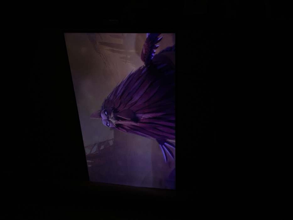 An image captured in Dobri dial, Bulgaria, depicts a widescreen projection in a darkened environment. The projection displays an animated, stylized avian creature with dark purple feathers, a prominent comb, a pointed beak, and glowing purple eyes set in a furrowed brow, conveying an aggressive or stern expression. The creature's body is largely comprised of long, layered dark purple plumage, with parts of its wings visible. The animated background features a desolate, textured landscape with dark, skeletal structures resembling a shipwreck on the left and multiple spire-like formations on the right, under a dark, transitionally colored sky. The immediate physical surroundings outside the projection are uniformly dark and featureless.