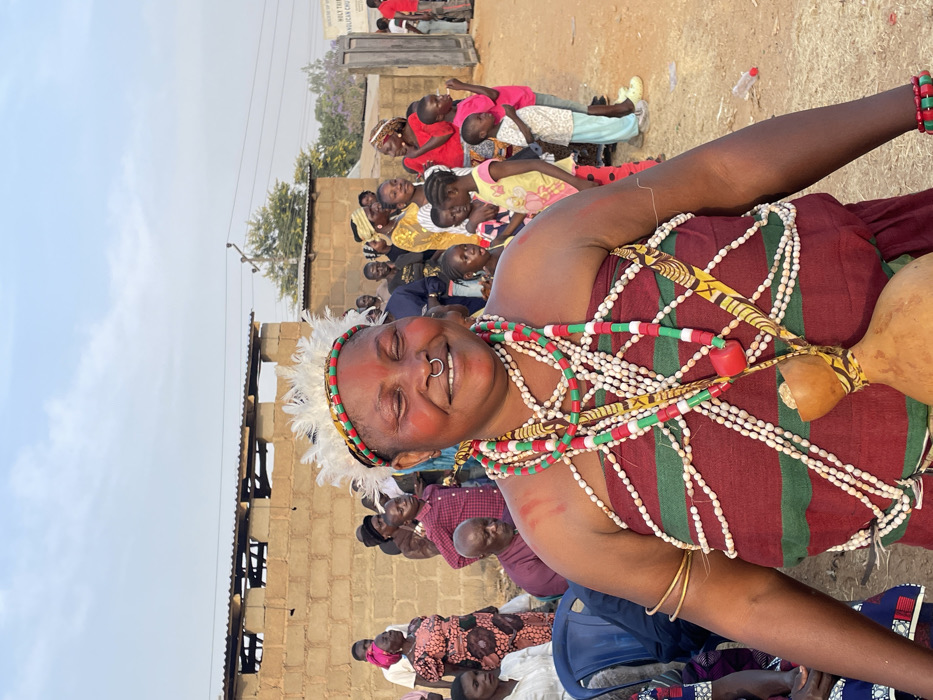 The image captures a vibrant outdoor gathering, with a focus on a smiling woman adorned in traditional attire. The photograph is oriented vertically, showing the woman prominently in the foreground.
The central figure is a woman with dark skin, broad features, and a joyful, wide smile. Her eyes are closed in her expression of happiness. She has red markings on her cheeks and a metal septum piercing in her nose. Her head is decorated with a white feathered headpiece secured by a band of red, green, and yellow patterns. She is heavily adorned with multiple strands of beads; some are small white beads, while others are larger red and green cylindrical beads, creating a rich, layered effect across her chest and shoulders. Her attire includes a fabric garment, possibly a wrapped cloth, featuring alternating wide stripes of dark red/maroon and thinner stripes of green with yellow patterns. A light brown gourd is visible on her right side, attached to her clothing. She wears simple gold-colored bangles on her left wrist and bracelets made of red and green beads on her right wrist.
Behind the woman, a crowd of people, including many children and adults, are gathered. They appear to be seated or standing, observing an event or performance. Several children are visible in the mid-ground, some looking in the same direction as the main subject, others glancing towards the camera. The setting appears to be a rural or semi-urban area, with simple buildings constructed from concrete blocks forming the backdrop. The ground is bare earth or dirt. In the upper right background, a faded sign is partially visible, on which the words "AN CHU" can be deciphered, likely part of "CHURCH". A tree with purple blossoms can be seen in the distance, and utility poles and wires crisscross against a light, clear sky, indicating daytime. The overall atmosphere conveyed by the main subject's expression is one of festivity and joy.