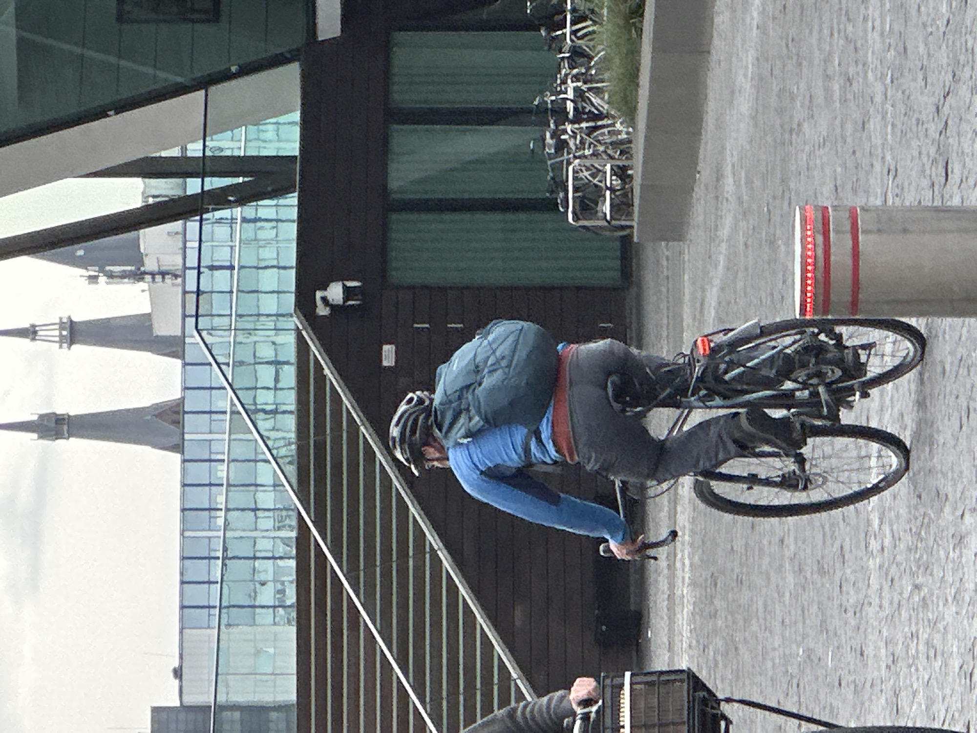 A cyclist wearing a blue long-sleeved shirt, gray pants, a helmet, and a backpack rides a bicycle on a cobblestone street. A second cyclist is partly visible in the lower foreground. A modern building with glass windows and metal accents is on the left. Parked bicycles are visible in the background. A cylindrical post with red and white stripes stands on the right. The scene is in Amsterdam, Netherlands, given the architectural style and the presence of numerous bicycles. A security camera is affixed to the building.