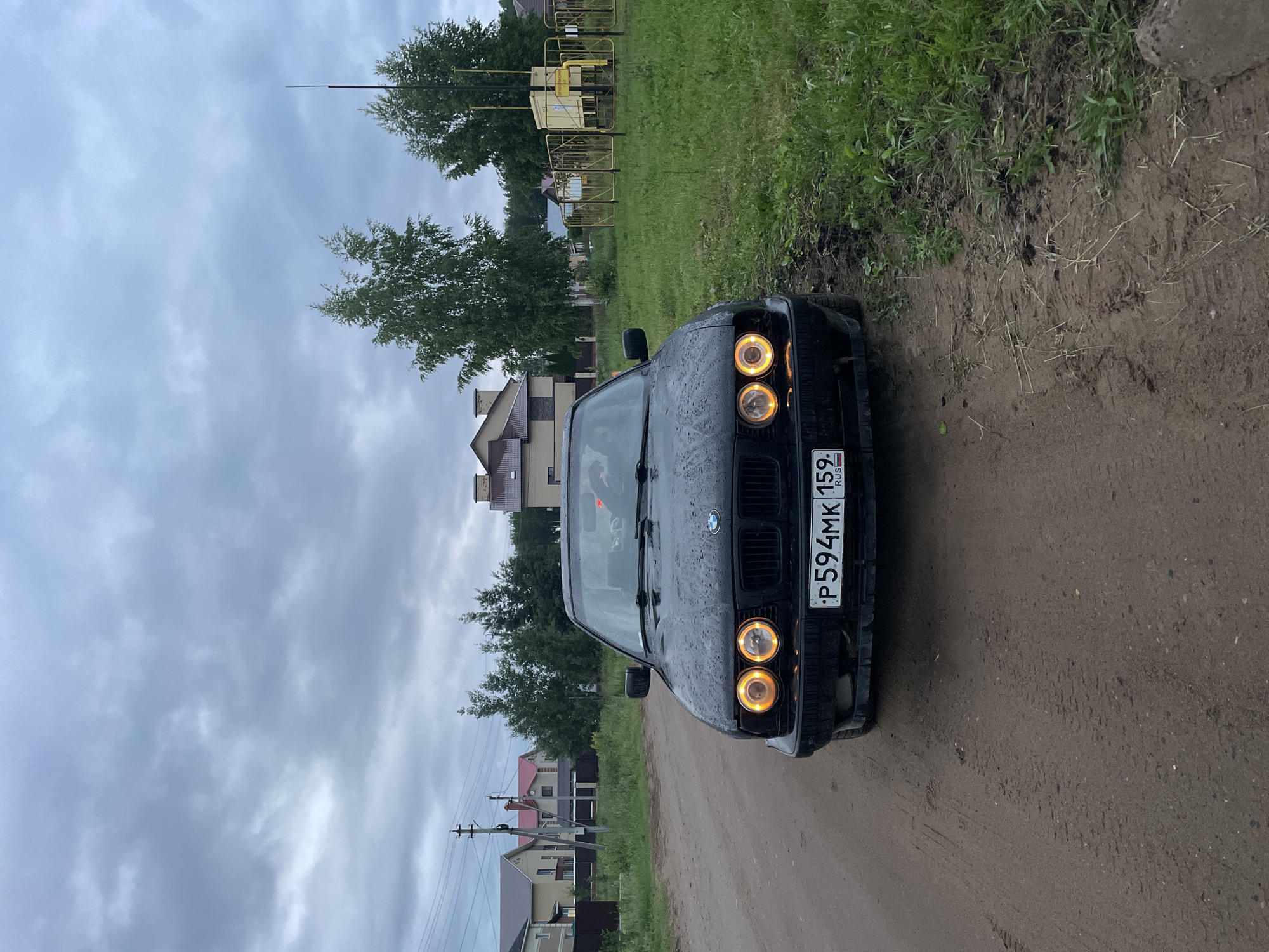 The image presents an outdoor scene captured on an overcast day in Noviy, Perm Krai, Russia. The primary subject is a black BMW sedan, likely an E34 5-series model, positioned centrally on an unpaved dirt or gravel road, facing the viewer. The vehicle's exterior, particularly the hood and windshield, is covered in visible water droplets, indicating recent precipitation. Its front headlights are illuminated, featuring distinct circular yellow-orange "angel eye" halo rings and inner lights. The front license plate, "P594MK 159 RUS," is clearly legible. Minor scuffing and wear are visible on the lower front bumper. A partial human figure is faintly discernible in the driver's seat, obscured by reflections on the windshield.
The road surface in the foreground and midground consists of damp, light brown dirt with visible tire tracks and ruts, suggesting a muddy condition. To the immediate right of the car, the ground transitions to sparse green grass.
In the background, on the left side of the road, a residential area is visible. Multiple modern, two-story houses with varying roof colors (brown and reddish-brown) are set back behind fences and sparse trees. Utility poles and overhead power lines run along the left side of the road.
On the right side of the image, beyond the grassy area, several deciduous trees with light-colored bark are prominent. Further back and to the right, a bright yellow metal structure, identified as a gas distribution or regulation station, is enclosed by a matching yellow fence. This station consists of interconnected yellow pipes and valves. A sign with Russian text, likely "ОГНЕОПАСНО" (Flammable), is attached to the equipment. A tall, slender, black pole or antenna-like structure rises vertically behind the gas station. The sky above is entirely covered by thick, grey clouds, indicating an overcast and potentially damp weather condition.