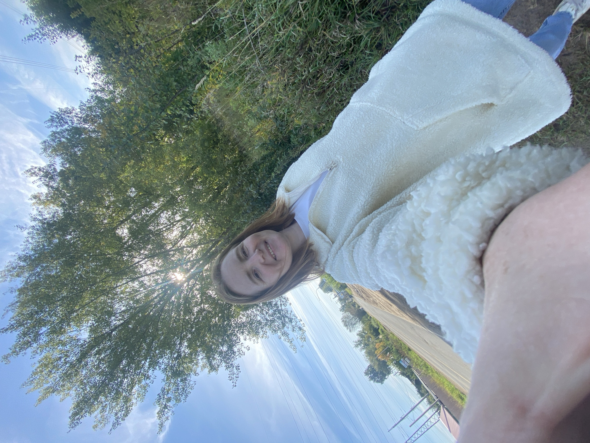 A woman is taking a selfie outdoors, near a road, with trees and grass in the background. She is wearing a cream-colored fleece jacket over a white t-shirt and blue jeans. The sun is shining brightly, casting a glow through the leaves of a large tree. The road appears to be in a rural or semi-rural area, with some houses visible in the distance. The sky is blue with some wispy white clouds. Power lines are visible in the upper part of the image. The location is identified as Chaikovsky, Russia.