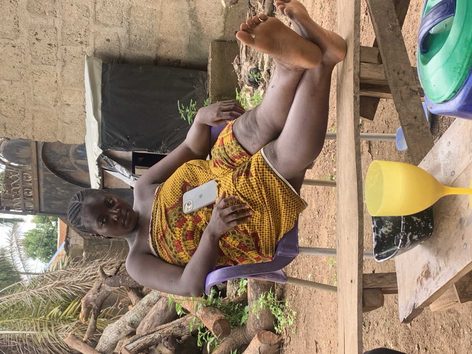 An outdoor, eye-level shot captures a woman reclining on a purple plastic chair in what appears to be a rustic yard in Akwanga, Nigeria. The woman, with braided hair and dark skin, is dressed in a vibrant yellow and red patterned fabric wrap. Her legs are crossed, with her bare feet visible and slightly raised. She holds a silver smartphone, characteristic of an iPhone, on her lap. She looks towards the camera with a neutral expression.
The setting includes a rough, light-colored concrete or mud brick wall in the background to the left. Below the wall, a black plastic sheet or tarp is visible. To the left of the woman, a pile of cut logs and branches, along with dry palm fronds and green ground cover, suggests an area with natural vegetation or discarded wood. To her right, a simple wooden table or bench holds a bright yellow plastic cup and a blue and green plastic container. The ground is dirt or sand. The scene is illuminated by natural light, indicating daytime. The overall impression is one of a relaxed, everyday moment in a rural or semi-urban environment. No specific text is clearly legible on any items.