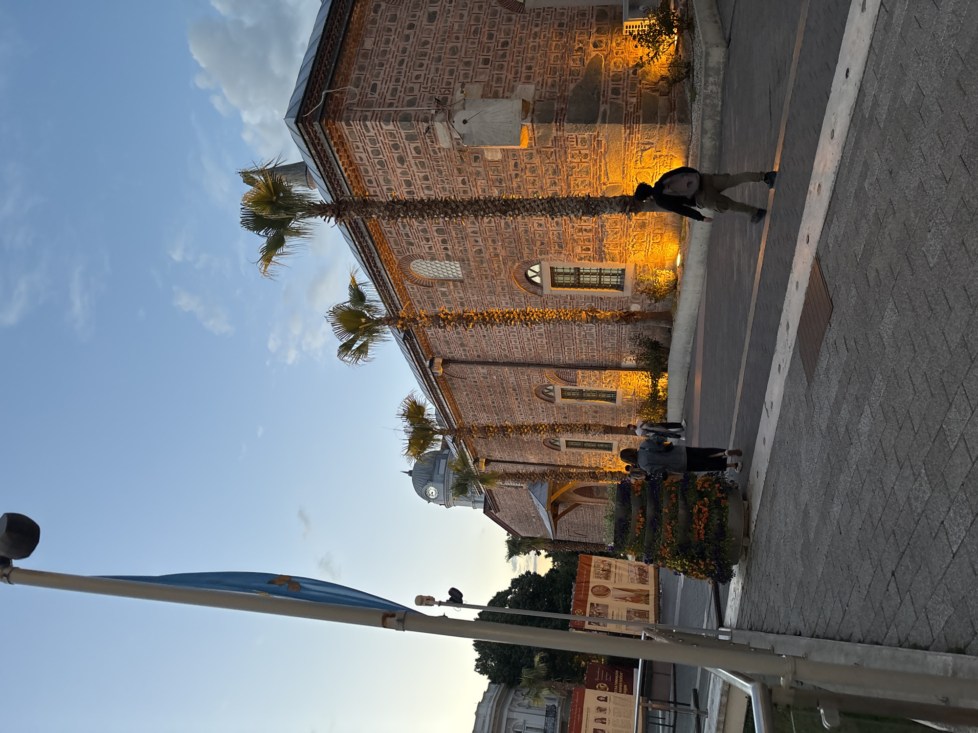 The image shows the Dzhumaya Mosque in Plovdiv, Bulgaria, taken at dusk. The building is made of brick and stone, with a sundial mounted on the exterior. Palm trees are planted in front of the mosque. People are walking on the sidewalk beside the building. A flagpole with a blue flag is visible in the foreground. A sign containing text and images is also visible along the sidewalk. The sky is a mix of blue and light clouds. Street lighting illuminates the mosque's facade. The ground is paved with patterned stones.