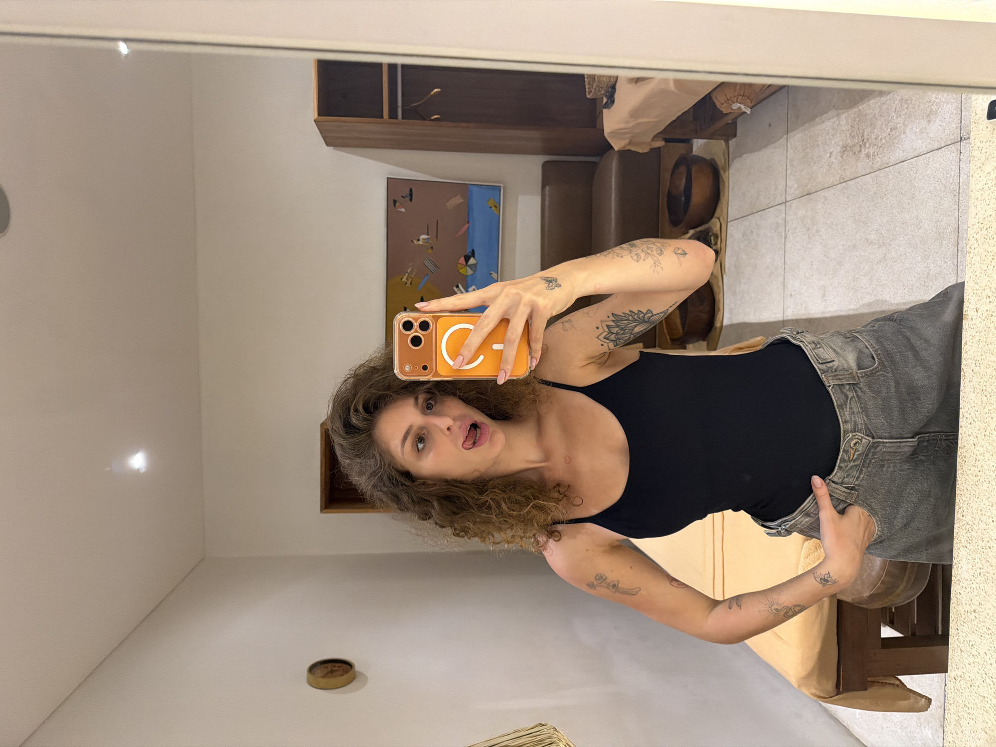 A young woman with curly brown hair stands in a room and takes a mirror selfie. She is wearing a black tank top and grey denim shorts. She has tattoos on her arms. Her phone, which has an orange case, is held up to take the photo. She is sticking her tongue out. The room appears to be a spa or massage room, with massage tables, wooden bowls, and a closet visible in the reflection of the mirror. The walls are white and the floor is tiled. A painting of people at a beach hangs on the wall behind her. The room is located in North Kuta, Indonesia.